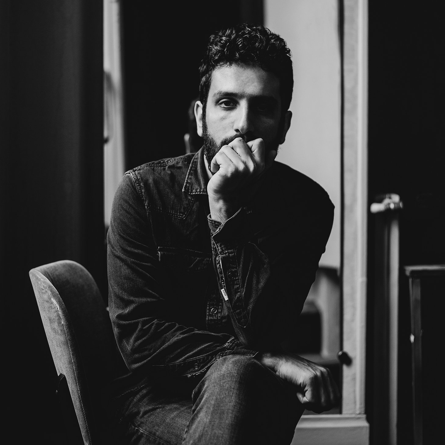 L'image montre un homme assis sur une chaise dans un intérieur sombre. Il a une barbe et porte une chemise en denim. Son expression est réfléchie, avec une main sur le menton, tandis qu'il regarde en direction de la caméra. L'éclairage met en valeur les contours de son visage et crée une ambiance introspective. Des éléments de l'environnement, comme une fenêtre et un miroir, ajoutent de la profondeur à la composition.