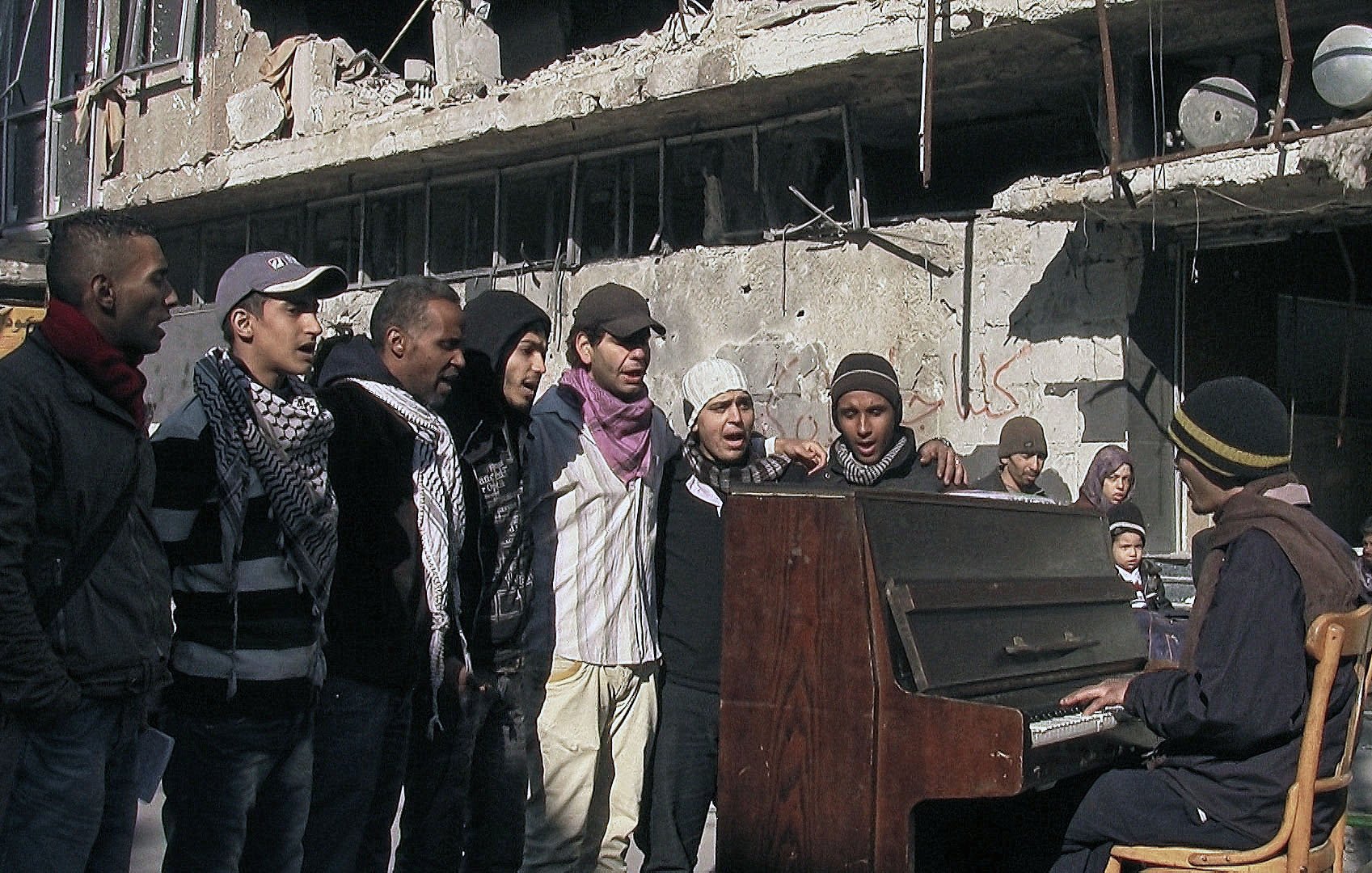 L'image montre un groupe de jeunes hommes chantant autour d'un piano dans un environnement dégradé, probablement après un conflit. Les bâtiments en arrière-plan sont en ruines, témoignant de destructions significatives. Les chanteurs semblent engagés et déterminés, tandis qu'un homme joue du piano, créant une atmosphère de solidarité et d'espoir au milieu de la désolation.