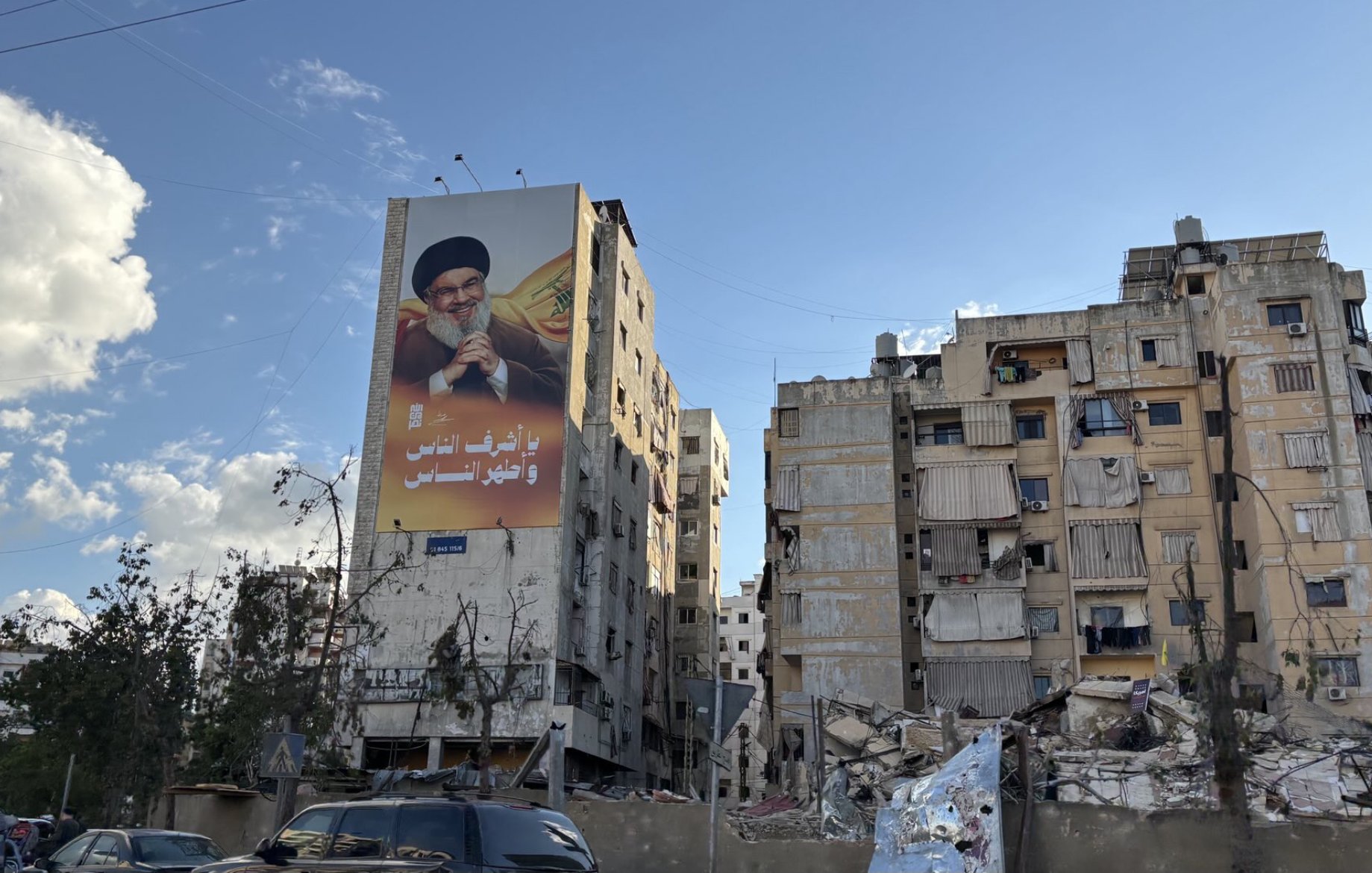 تظهر الصورة مبنى متهدم جزئيًا في منطقة حضرية، حيث تتواجد فيه علامات على الدمار. في الجهة العليا من المبنى، يوجد إعلان كبير يحمل صورة لشخصية معروفة، وعليه نص باللغة العربية. في الخلفية، تظهر السحب وبعض الأشجار المتضررة. المنظر يعكس أجواء من التوتر والدمار، مما يشير إلى حالة من الأزمات أو الصراعات في المنطقة.