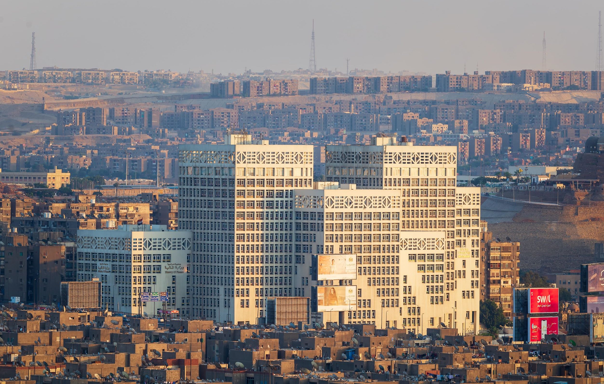 تظهر في الصورة مدينة تتميز بكثافة المباني والشقق السكنية. هناك مجموعة من المباني الحديثة ذات التصميم الفريد في المقدمة، حيث ينعكس عليها ضوء الشمس مما يبرز تفاصيلها. خلفها، تظهر منطقة سكنية كثيفة البناء، مع مشهد لجوانب مختلفة من المدينة. يبدو أن الجو هادئ، مع سماء مضيئة وقليل من الضباب.