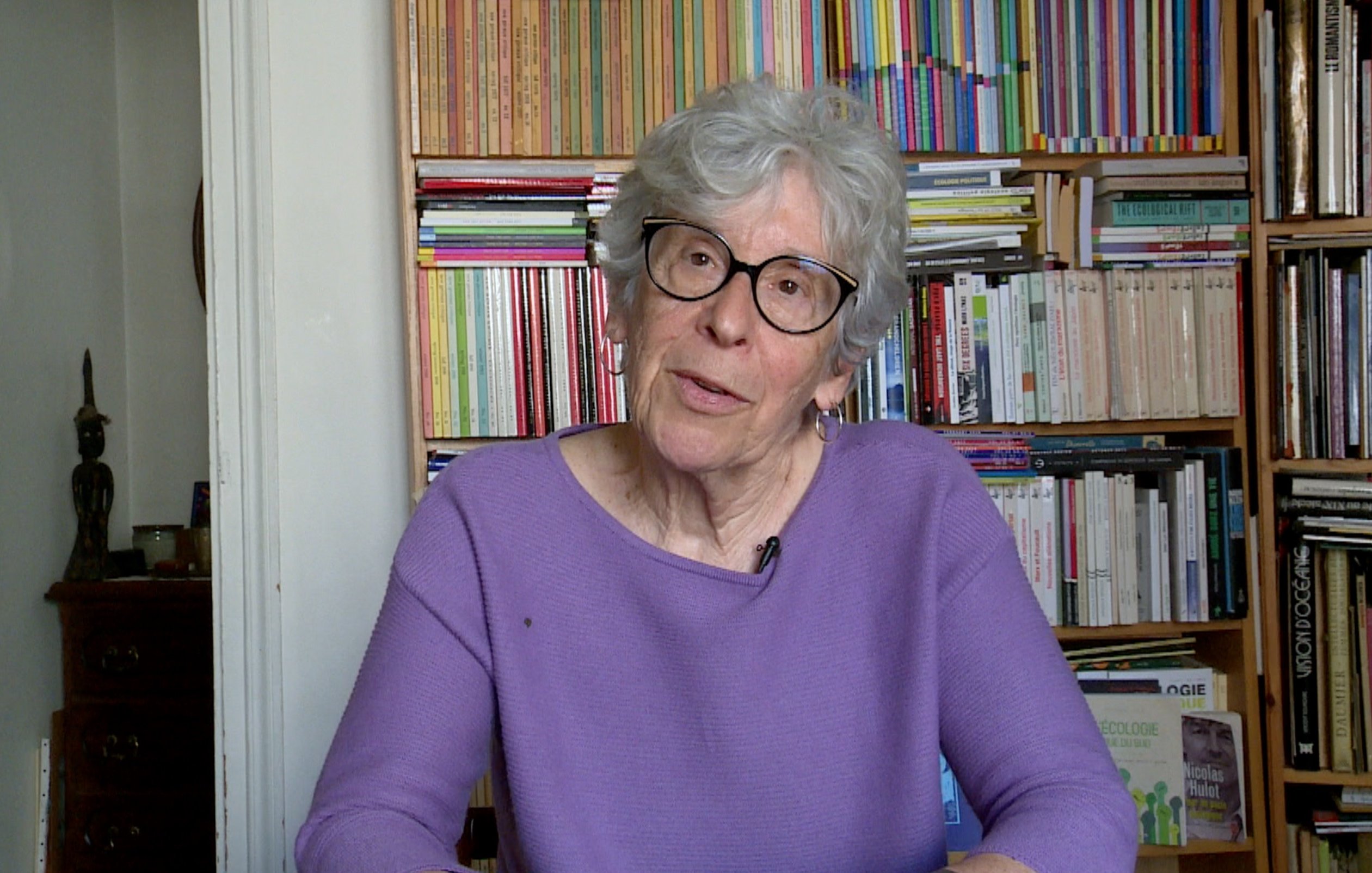 L'image montre une femme âgée avec des cheveux gris et des lunettes. Elle porte un pull violet et est assise devant une bibliothèque remplie de livres, qui présentent des couvertures colorées. Son expression est réfléchie et elle semble engagée dans une conversation. L'environnement est chaleureux et intellectuel, avec des étagères bien ordonnées autour d'elle.