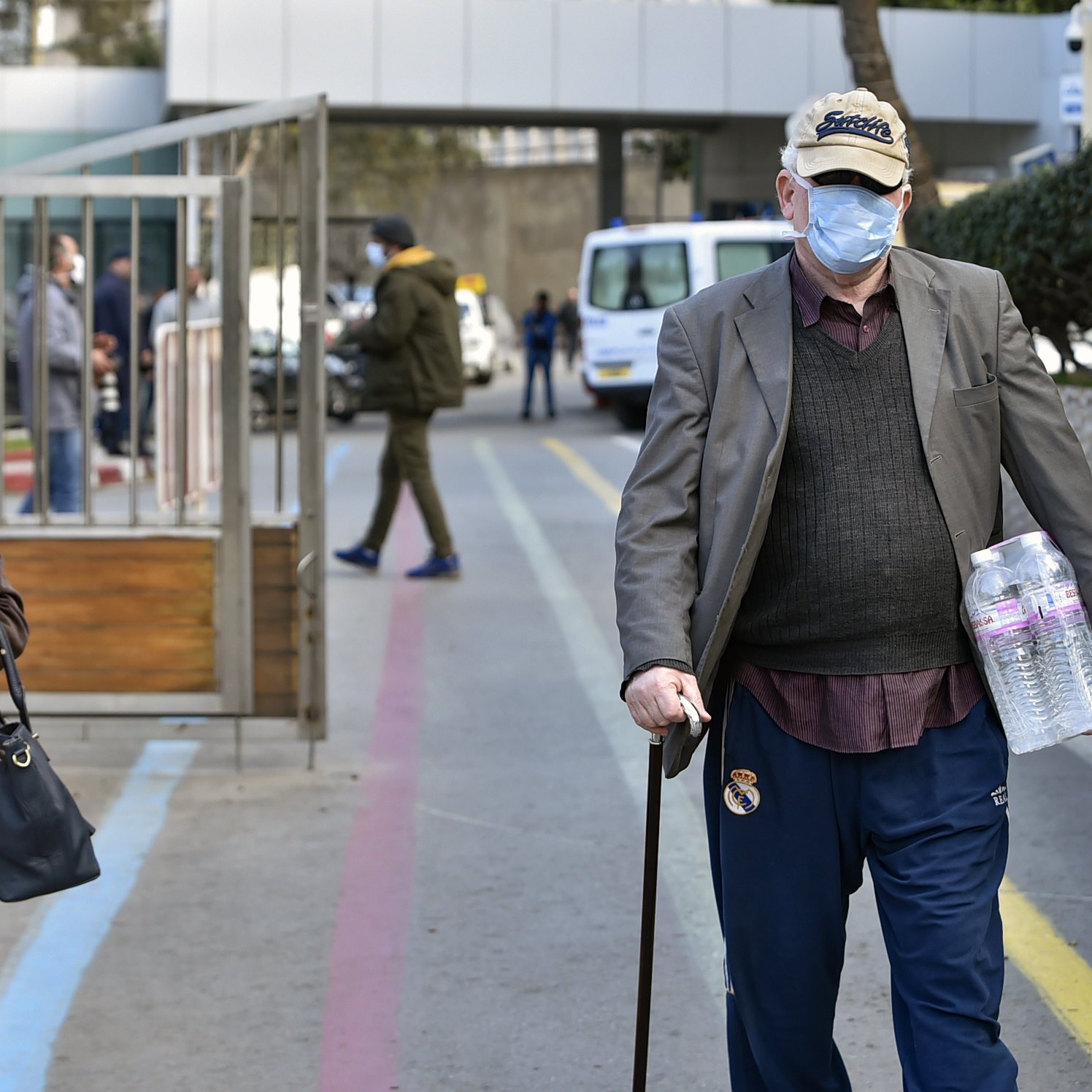 L'image montre une scène urbaine où deux personnes marchent dans une rue. À gauche, une femme portant un foulard et une veste rouge tient un sac, tandis qu'à droite, un homme âgé, avec une canne et un chapeau, marche en portant deux bouteilles d'eau. Les deux portent des masques faciaux, suggérant un contexte sanitaire. En arrière-plan, on aperçoit des véhicules et des bâtiments.