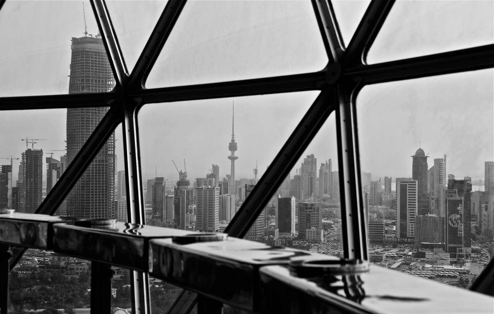 L'image montre une vue urbaine à travers une grande fenêtre en verre avec un cadre géométrique. Au premier plan, on aperçoit une surface réfléchissante et des éléments architecturaux. À l'extérieur, on découvre un paysage de ville en noir et blanc, avec des gratte-ciels modernes et une tour emblématique qui se dresse au loin, probablement un symbole de la skyline. La scène dégage une atmosphère à la fois contemporaine et dynamique.