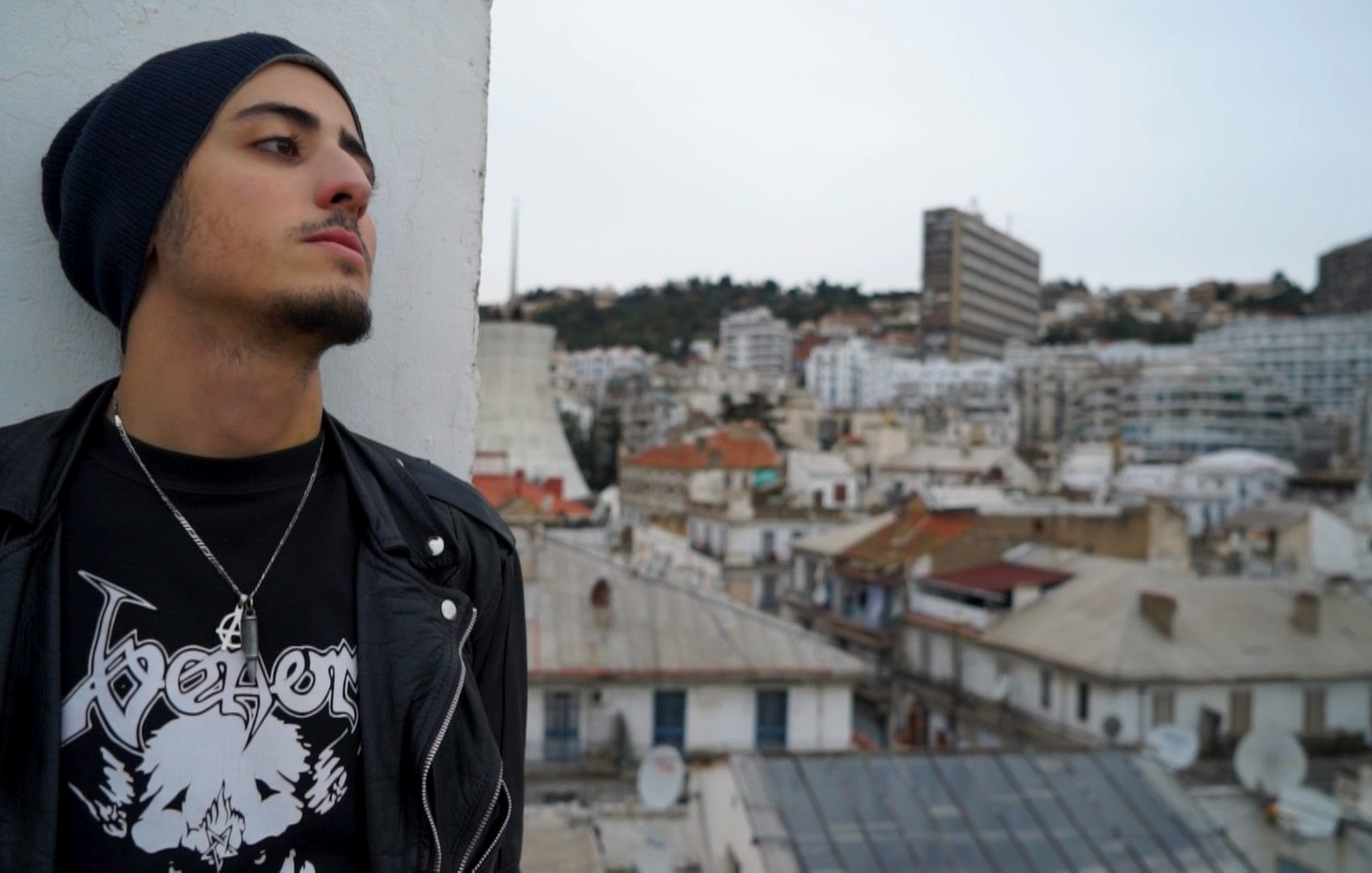 L'image montre un jeune homme qui se tient sur un toit, regardant au loin. Il porte un bonnet et une veste en cuir noire, ainsi qu'un t-shirt avec un design graphique. En arrière-plan, on aperçoit une ville avec des bâtiments de différentes tailles et architectures, et des toits de maisons. Le ciel est légèrement nuageux, ce qui donne une ambiance calme et contemplative à la scène.