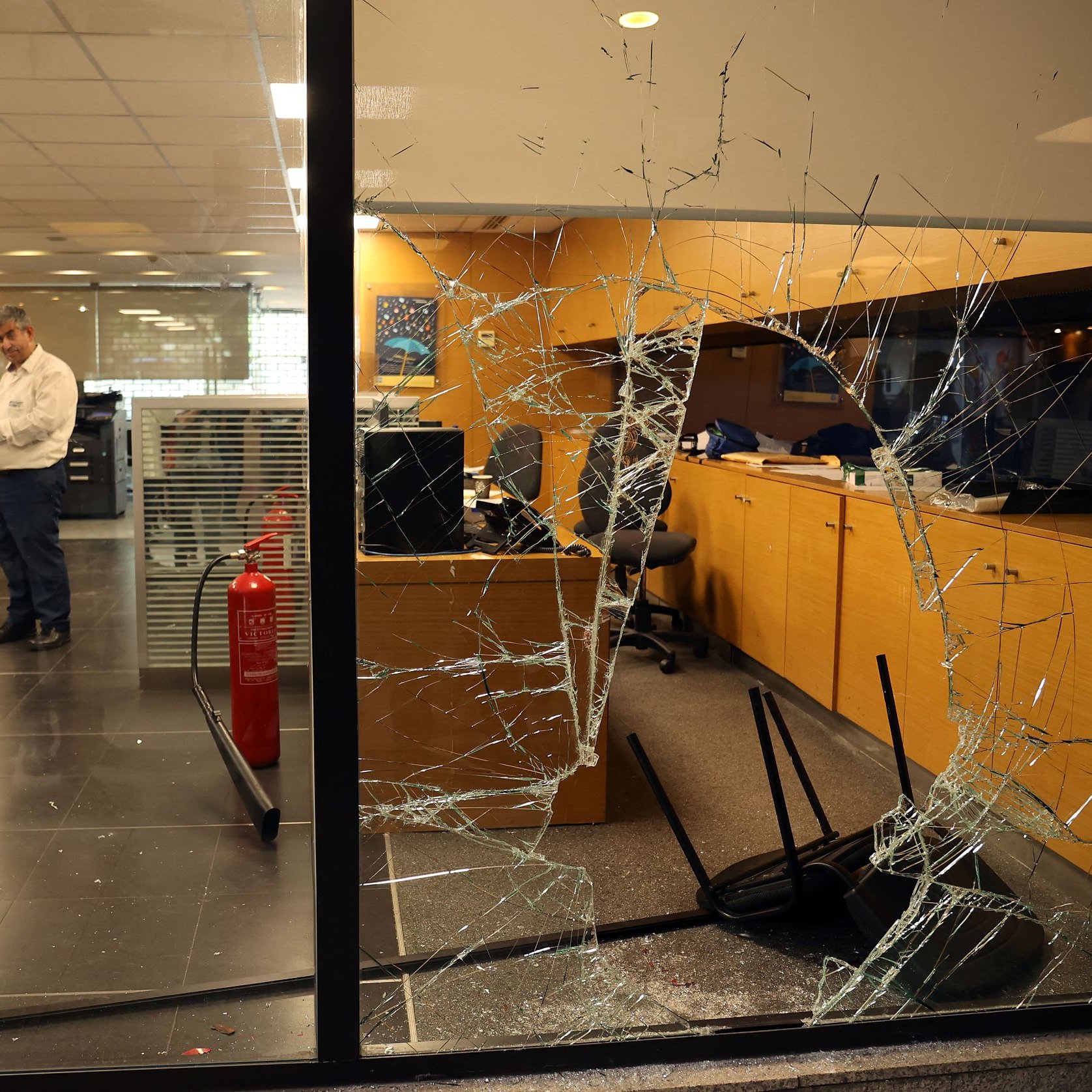 L'image montre un bureau avec des dégâts visibles. On peut voir une vitre brisée à l'avant, avec des fissures qui s'étendent sur toute sa surface. À l'intérieur, un homme est debout, semblant examiner la situation, tandis que des chaises sont renversées sur le sol. L'ambiance générale suggère qu'il y a eu un incident perturbateur dans cet espace de travail.