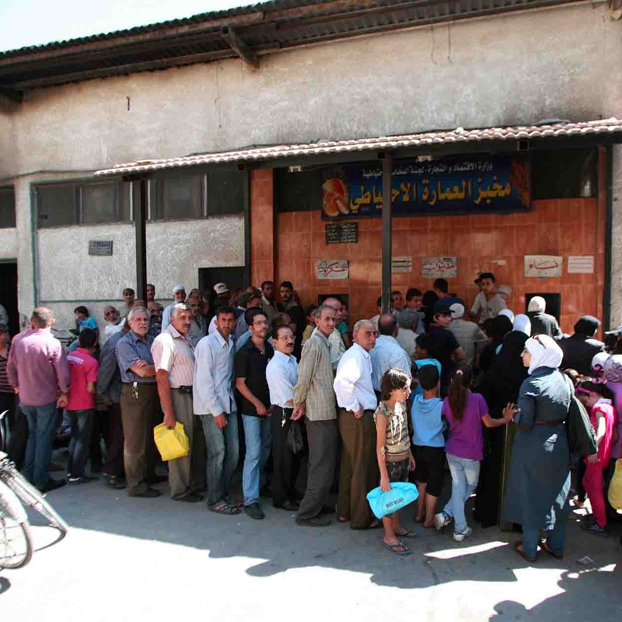 الصورة تظهر تجمعاً كبيراً من الناس أمام مبنى يبدو أنه متجر أو مركز خدمات. تتنوع أعمار الأفراد في الصف، حيث يوجد رجال ونساء، وبعضهم يحمل حقائب أو أكياس. المبنى يظهر بواجهة بسيطة، وهناك لافتة مكتوبة باللغة العربية. الأجواء تبدو نشطة، مع وجود أشخاص يراقبون أو يتحدثون مع بعضهم البعض. الخلفية تحتوي على عناصر حضرية مثل الدراجات والعمارات.