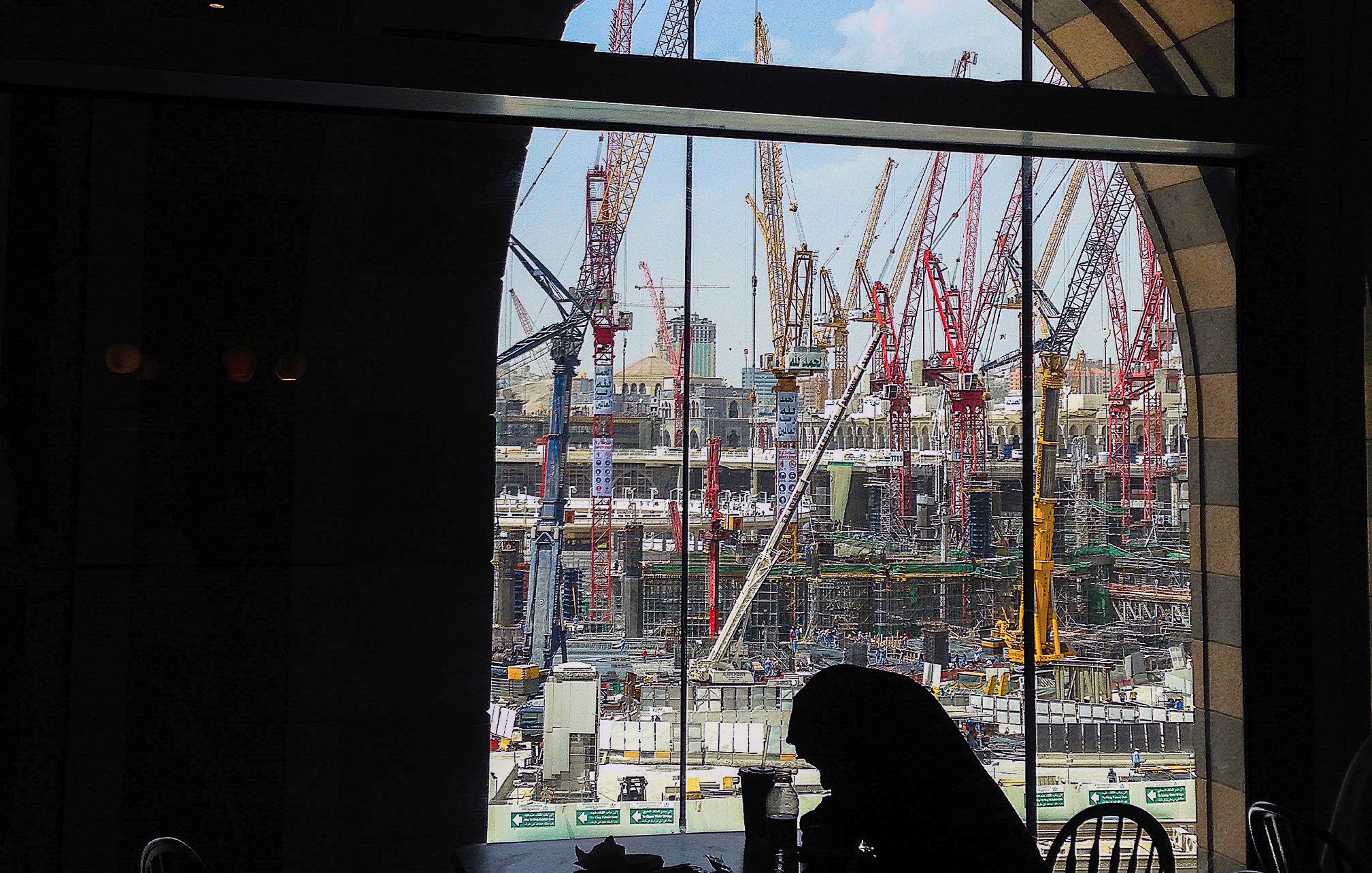 L'image montre un chantier de construction vu à travers une grande fenêtre à arc. À l'intérieur, on aperçoit une silhouette assise à une table. De l'autre côté de la vitre, de nombreux grues et engins de chantier se dressent, travaillant à un projet de grande envergure. Le ciel est partiellement dégagé, ajoutant une touche de lumière à la scène. L'ensemble dégage une atmosphère de travail intensif et de développement urbain.