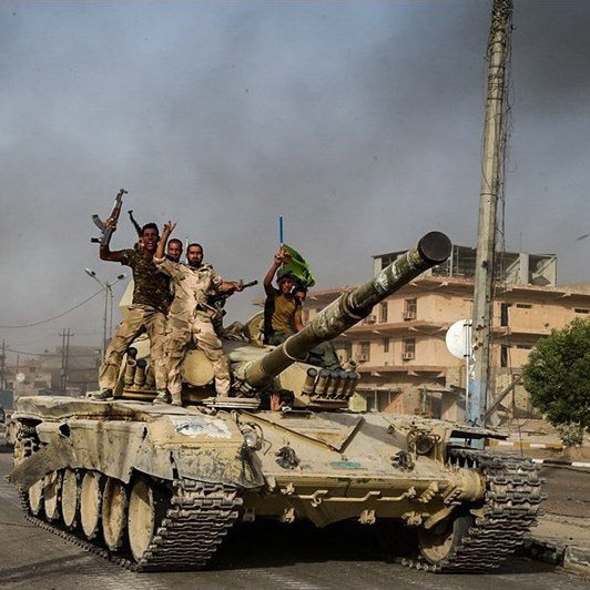 L'image montre des tanks avancant sur une route dans un environnement urbain. Plusieurs soldats sont visibles sur les tanks, certains levant les bras en signe de victoire ou de célébration. L'arrière-plan présente des bâtiments endommagés et une atmosphère chargée de fumée, ce qui suggère un contexte de conflit. Les couleurs sont principalement terreuses, avec des teintes de vert et de beige des tanks et des uniformes des soldats.