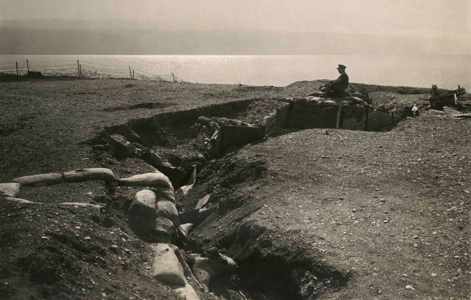 L'image montre une tranchée militaire dans un paysage aride. On peut voir une personne assise sur le sommet d'un rempart en sable, observant les environs. La tranchée est entourée de sacs de sable, et l'horizon est marqué par un plan d'eau au loin. L'atmosphère est sombre et pourrait évoquer une période de conflit, comme la Première ou la Seconde Guerre mondiale. L'éclairage donne à la scène un aspect nostalgique et témoin de l'histoire.