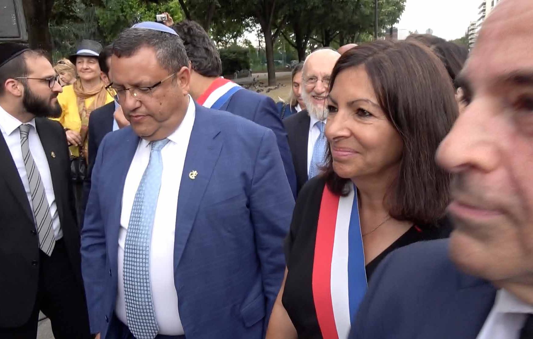 The image shows a group of people outdoors, likely at a public event. In the foreground, two individuals are prominently featured: one man is wearing a blue suit, and the other, a woman, is wearing a black outfit adorned with a ribbon that signifies a formal position. Both appear to be engaged in conversation or observing their surroundings. Other individuals are in the background, suggesting a social or official gathering. The setting appears to be a park or an outdoor area with trees.