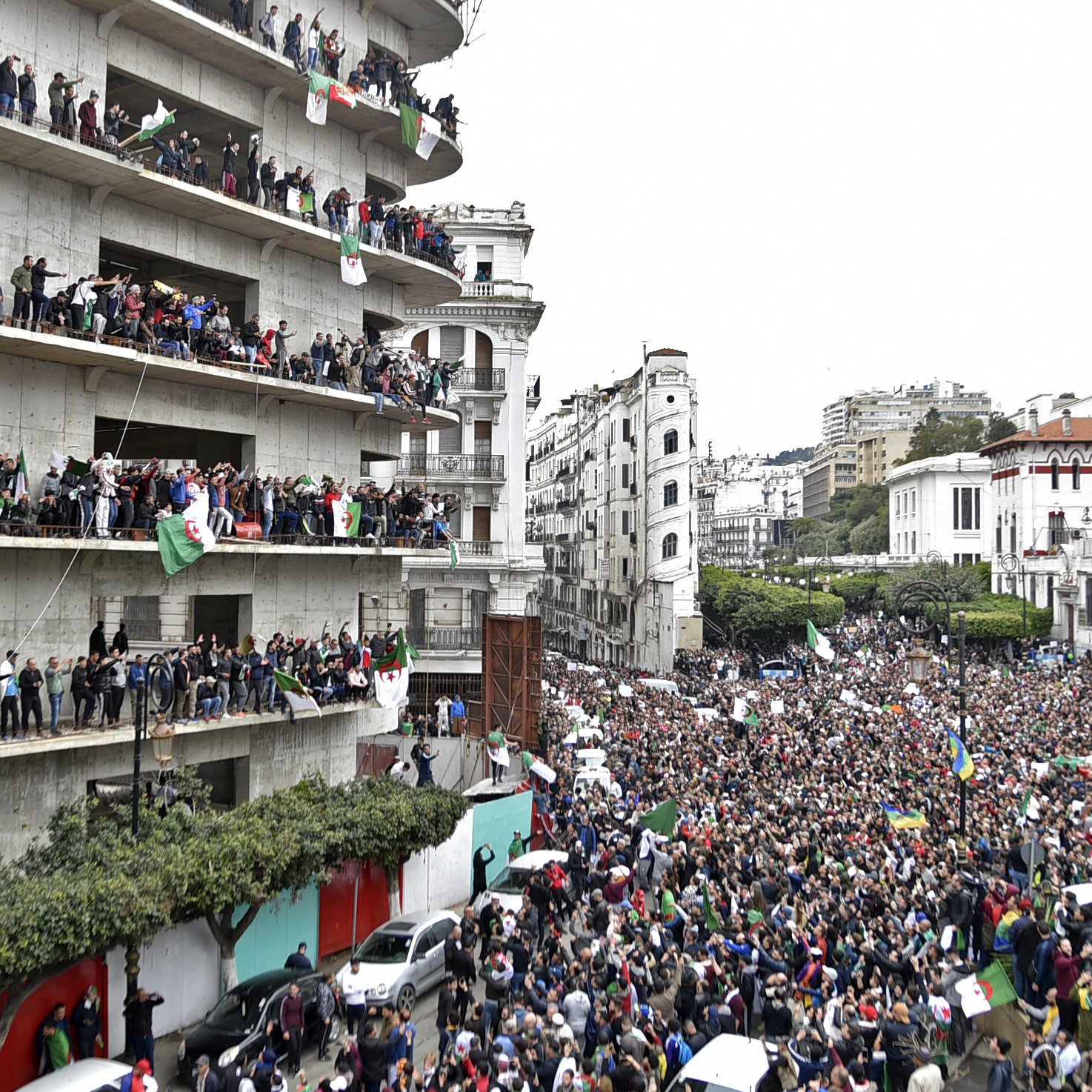 L'image montre une grande foule rassemblée devant un bâtiment, manifestant dans une ville. De nombreuses personnes sont visibles sur les balcons du bâtiment, brandissant des drapeaux et exprimant leur soutien à la manifestation. La rue est remplie de manifestants, et l'atmosphère semble vibrante et dynamique, avec un mélange de drapeaux et de pancartes. Le paysage urbain environnant, composé de divers bâtiments, ajoute à l'environnement de la scène.