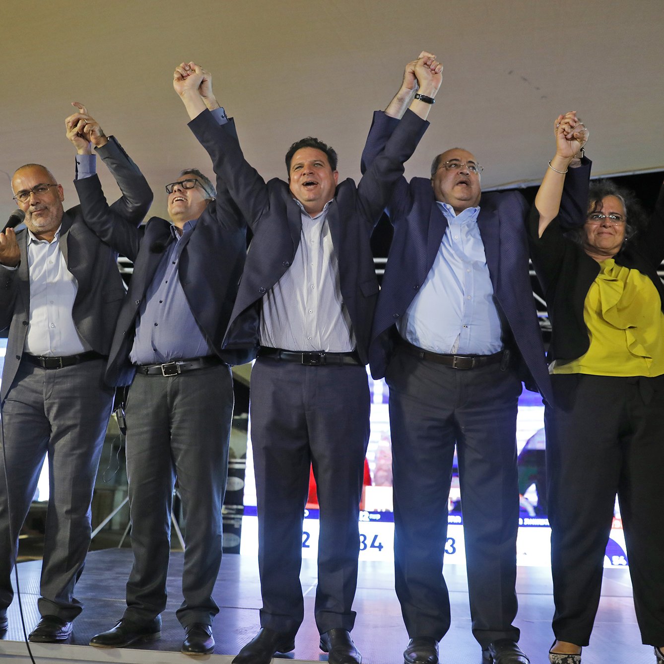 L'image montre un groupe de personnes sur une scène, levant les bras en l'air dans un geste de célébration ou de triomphe. Ils semblent être réunis pour un événement politique, probablement après l'annonce de résultats électoraux. Les participants portent des vêtements formels, et l'ambiance paraît festive et unie. En arrière-plan, on peut voir des écrans avec des informations, ce qui suggère que c'est une occasion importante.