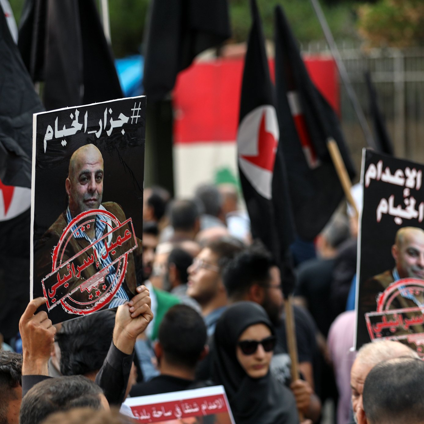 The image depicts a crowd of protesters holding signs and flags. The signs feature a portrait of a man along with a hashtag and phrases in Arabic, indicating a political or social issue. The crowd appears engaged and is likely part of a demonstration. The flags are predominantly black with red symbols, contributing to the atmosphere of the protest. The environment suggests a public gathering, possibly in response to a recent event or issue.