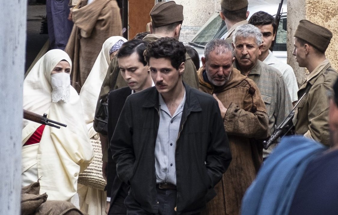 L'image montre une scène urbaine où plusieurs personnes sont rassemblées. Au centre, on voit un jeune homme en chemise claire et veste sombre, l'air pensif. Il est entouré de groupes de personnes, dont certaines semblent discuter ou se déplacer. Il y a également des hommes en uniformes militaires et des femmes portant des vêtements traditionnels, y compris un couvre-chef. L'ambiance de la scène semble chargée d'émotion, et le décor évoque une atmosphère historique ou dramatique.