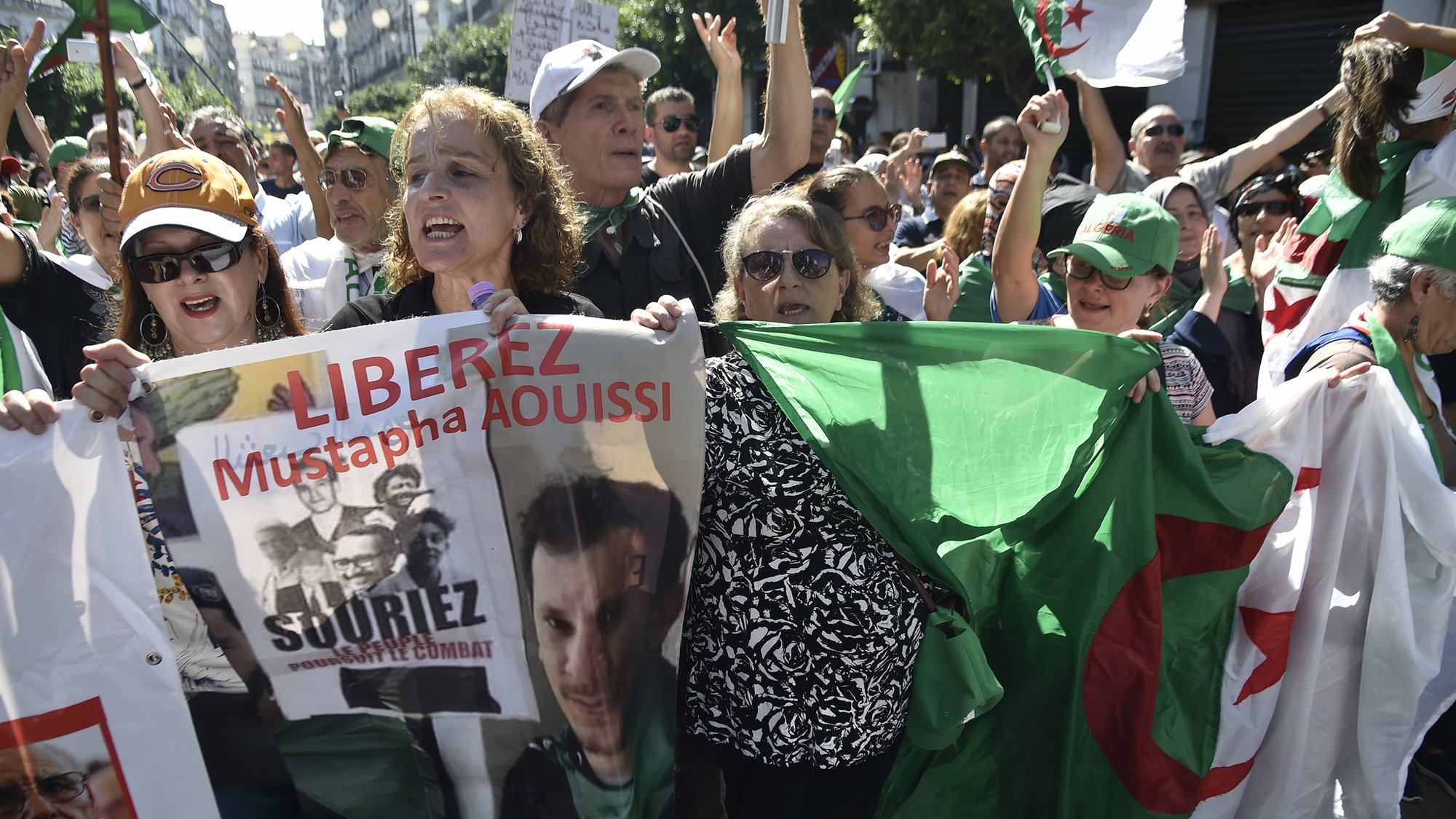 الصورة تظهر مجموعة من المتظاهرين في الجزائر، يحملون لافتات وأعلام. يبدو أن هناك نساء ورجال في المقدمة، ويعبرون عن مطالبات سياسية. إحدى اللافتات تحمل اسم "مصطفى عويصي" وعبارة "حرروا". الأجواء توحي بالحماسة والمطالبة بالتغيير.