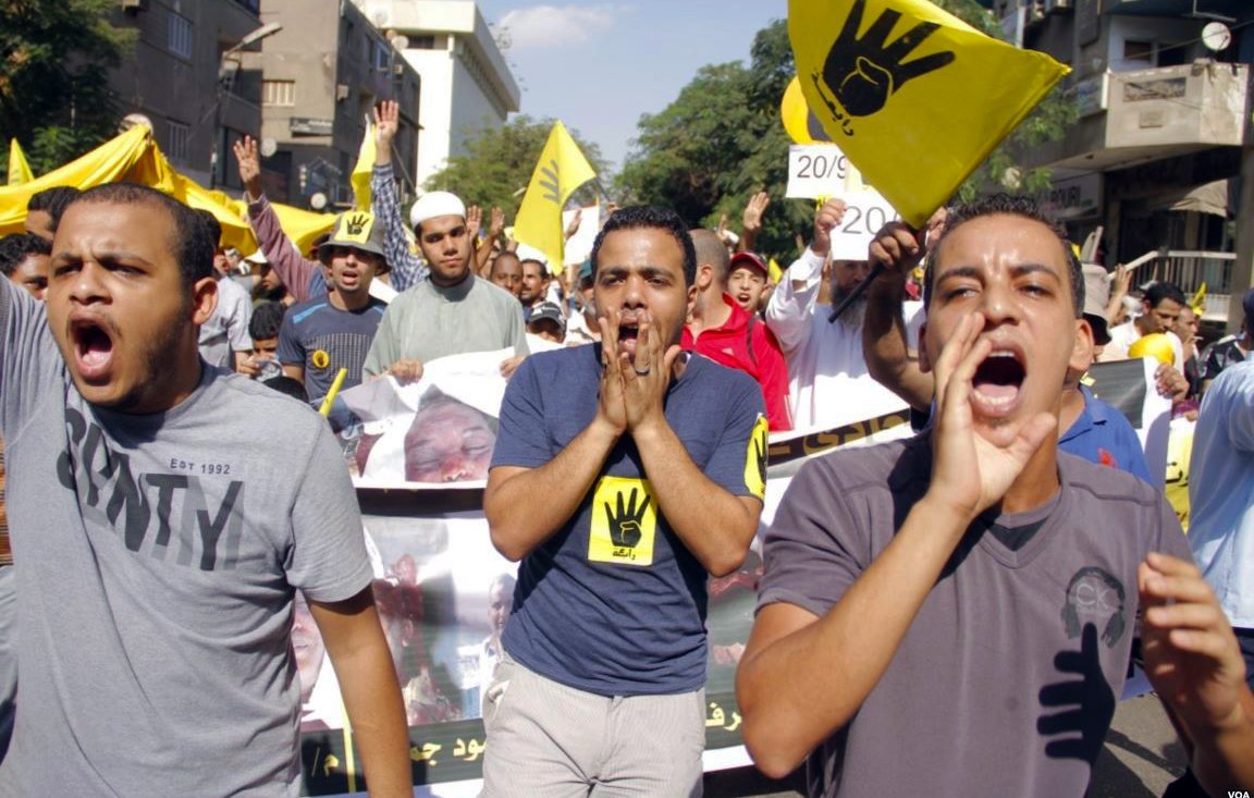 La imagen muestra a un grupo de personas participando en una manifestación. Los manifestantes parecen estar expresando su descontento, gritando y levantando las manos. En el fondo, se pueden ver pancartas y banderas, algunas de las cuales tienen un símbolo de mano levantada. El ambiente es de protesta, y la multitud está activa y unida en su mensaje. La escena se desarrolla en un entorno urbano, con edificios visibles en el fondo.