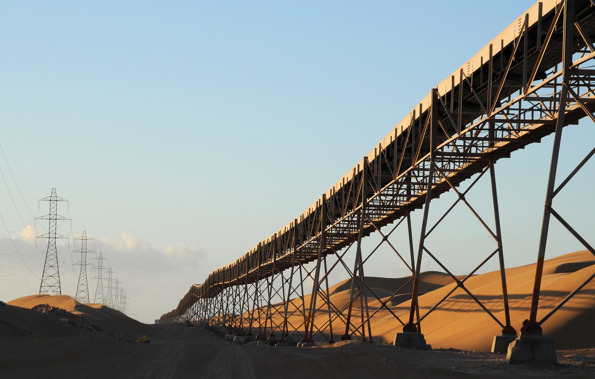 The image depicts a long conveyor belt system stretching across a vast landscape dominated by sand dunes. The conveyor belt is elevated on metal supports, running parallel to the ground. In the background, tall power lines extend into the horizon, hinting at an industrial setting. The warm light of the sun creates a golden hue on the sand, adding to the serene yet striking composition of the scene. The overall atmosphere suggests a remote, arid environment that may be associated with mining or agricultural operations.