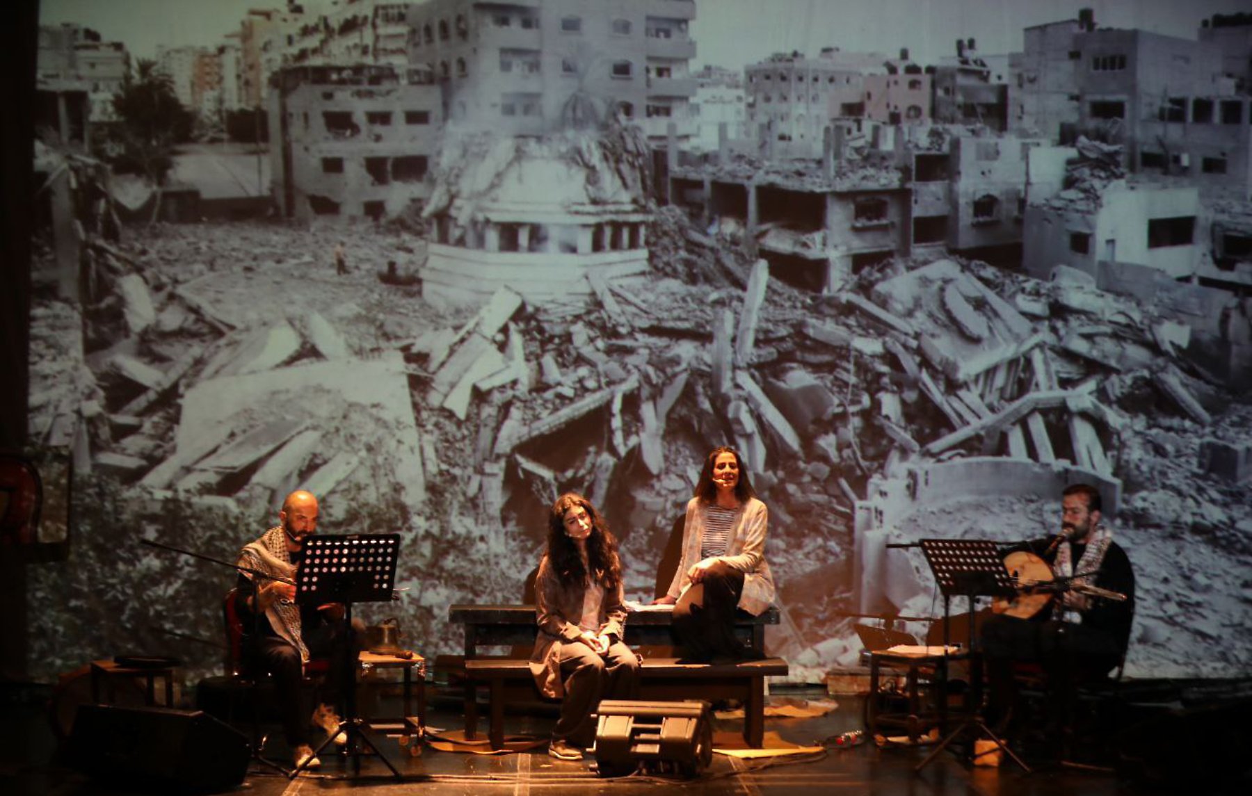 Four performers sit with instruments in front of a large backdrop of war-torn buildings.