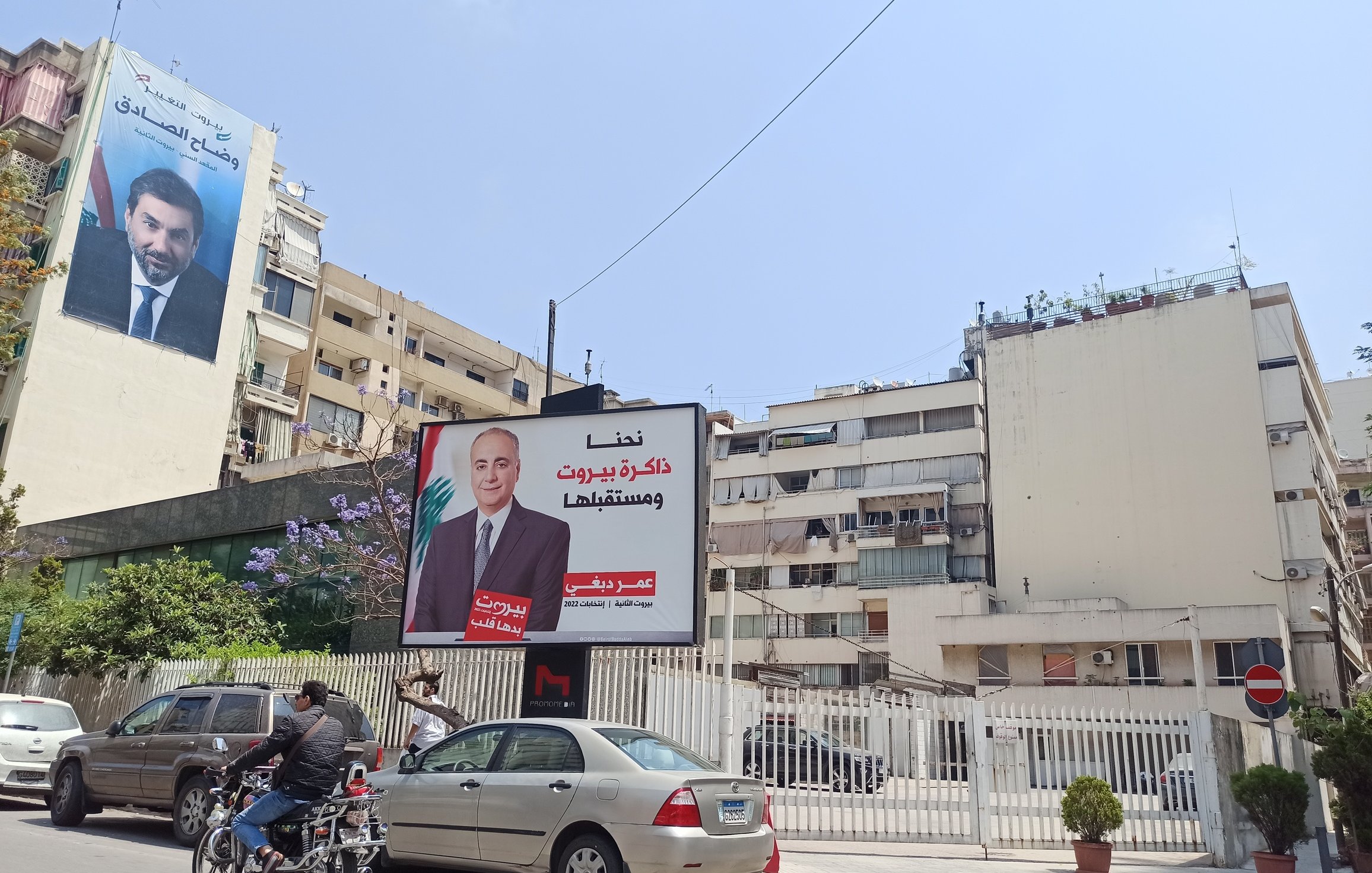 L'immagine mostra una scena urbana con un edificio signorile e un grande cartellone pubblicitario. Il cartellone presenta un ritratto di un uomo in giacca e cravatta, accompagnato da un testo in arabo. Accanto, si può notare un altro edificio che ospita un altro cartellone, ma parte del suo contenuto non è leggibile. Ci sono anche alcune automobili parcheggiate e un motociclista che passa. La via sembra essere molto luminosa grazie al sole.