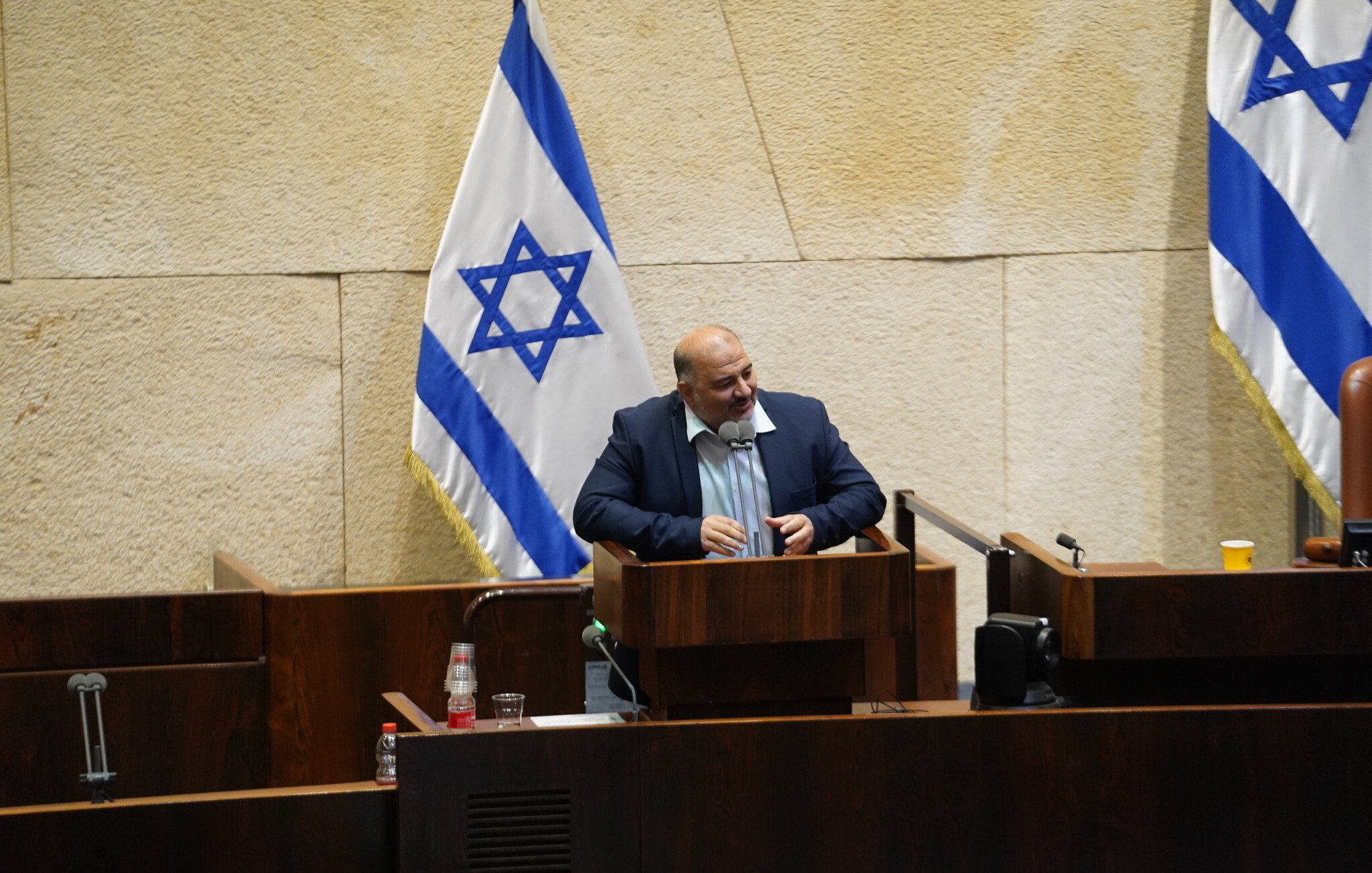 L'image montre un homme debout à un pupitre dans une salle de réunion, probablement au sein d'une assemblée ou d'un parlement, avec des drapeaux israéliens en arrière-plan. Le décor est sobre, avec des murs en pierre et du mobilier en bois. L'homme semble s'adresser à un public, exprimant peut-être son opinion sur un sujet d'actualité.