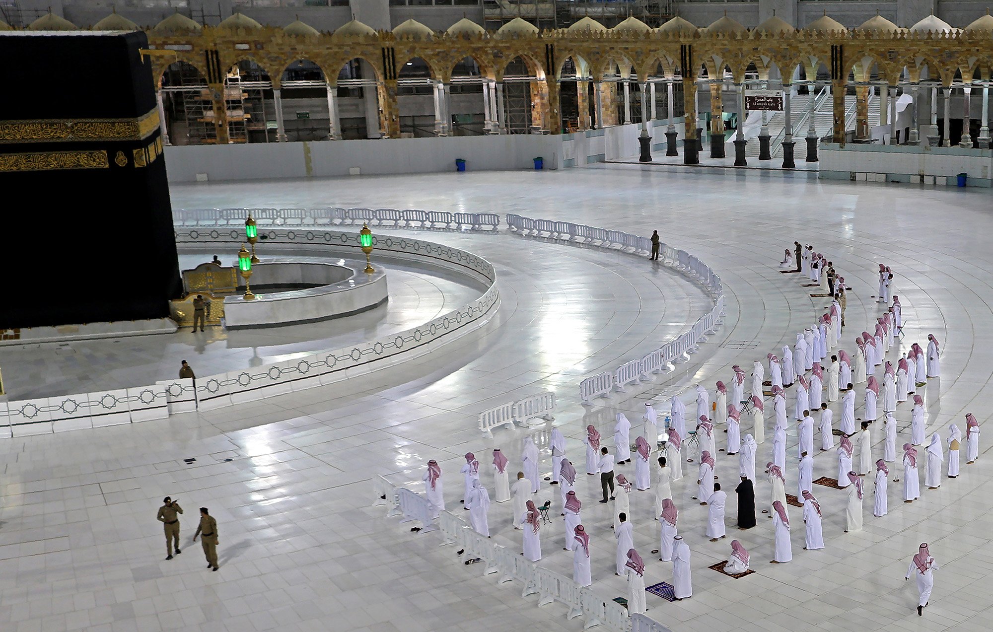 La imagen muestra el interior de la Gran Mezquita de La Meca, donde se encuentra la Kaaba. Se puede ver un gran espacio blanco con filas de personas en posición de oración. Están vistiendo tradicionalmente con túnicas blancas y pañuelos en la cabeza. En el fondo, se aprecia la Kaaba con su cubierta negra y algunos elementos decorativos. También hay barreras que delimitan el espacio y un par de agentes de seguridad en el área. La atmósfera es de calma y devoción.