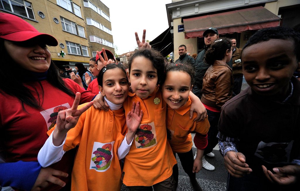 L'image montre un groupe d'enfants souriants, posant ensemble dans une rue animée. Ils portent des t-shirts colorés, principalement orange, avec des motifs ou des logos visibles. Les enfants font des signes de paix avec leurs doigts et affichent des expressions joyeuses. En arrière-plan, on aperçoit des adultes et d'autres participants à un événement festif. L'atmosphère semble conviviale et festive.