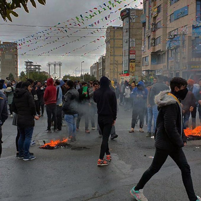 L'image montre une foule rassemblée dans une rue, probablement lors d'une manifestation. Plusieurs personnes portent des vêtements chauds et des masques. On peut observer des petits feux sur la chaussée, entourés de fumée. L'atmosphère semble tendue, avec des bâtiments et des décorations en arrière-plan. Le ciel est nuageux, ajoutant à l'ambiance sombre de la scène.