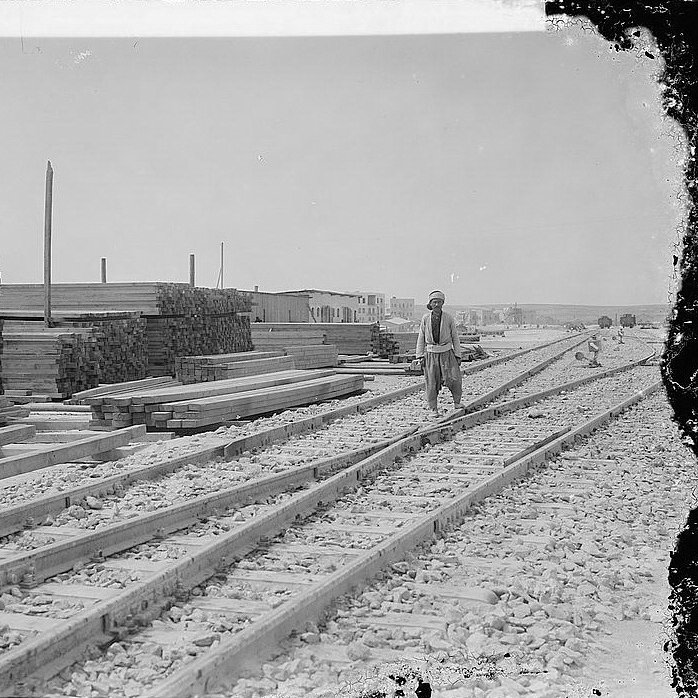 L'image montre une scène industrielle avec des voies de chemin de fer se dirigeant vers l'horizon. Sur le côté gauche, il y a des tas de planches de bois empilées, ce qui suggère une activité de construction ou de stockage. Un homme, probablement un travailleur, marche le long des rails. En arrière-plan, on aperçoit des bâtiments, donnant l'impression d'un site en pleine activité. Le ciel est dégagé, et la lumière est vive, typique d'une journée ensoleillée. L'atmosphère générale évoque une époque passée, avec une ambiance de travail acharné.