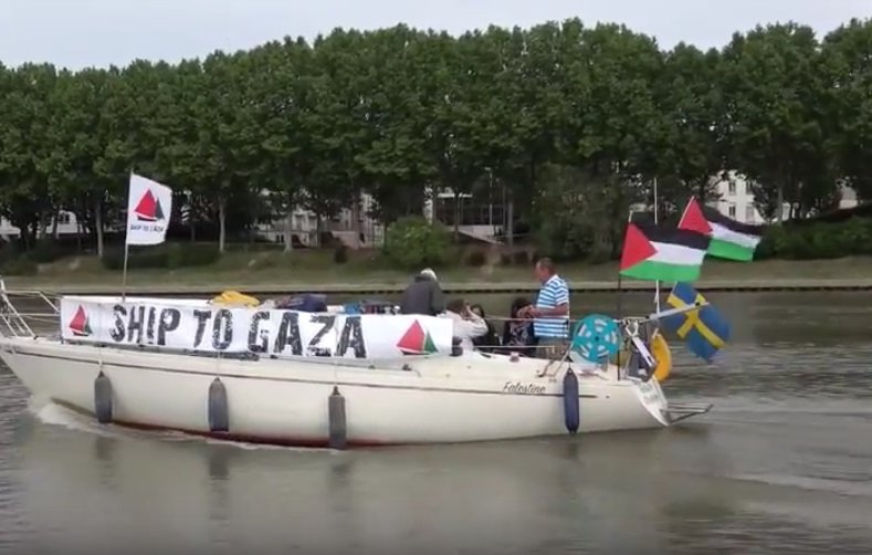 The image shows a boat on a body of water with several individuals on board. The boat has a prominent banner that reads "SHIP TO GAZA," and it is adorned with various flags, including the Palestinian flag and the flag of Sweden. In the background, there are trees and buildings along the shoreline. The scene conveys themes of solidarity and activism related to Gaza.