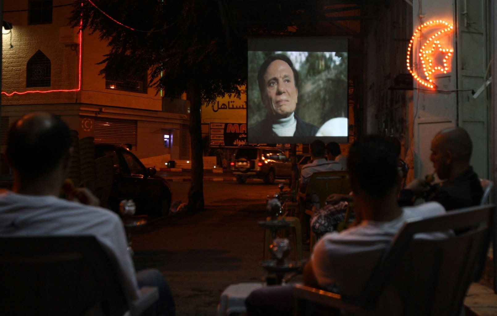 L'image montre une scène nocturne où un groupe de personnes est assis à l'extérieur, observant un film projeté sur un mur. On voit des chaises disposées face à l'écran, et quelques fumées de narguilé flottent dans l'air. L'environnement est animé par des lumières de néon, et on aperçoit des voitures garées et des bâtiments en arrière-plan, créant une ambiance conviviale et détendue.