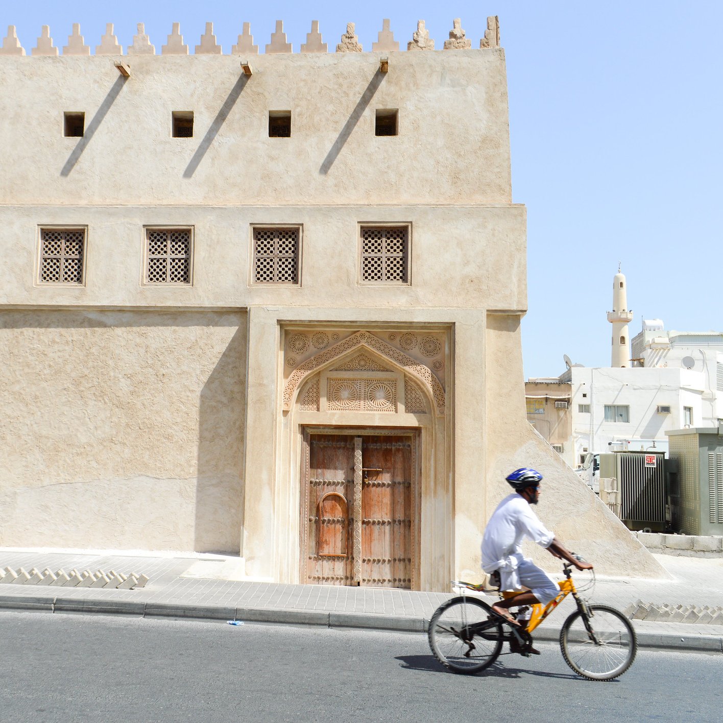 L'image montre un homme à vélo qui passe à côté d'un bâtiment au style architectural traditionnel. Ce bâtiment, de couleur sable, présente des murs texturés et des fenêtres ornées de motifs. On peut apercevoir une grande porte en bois sculpté, typique de l'architecture ancienne. En arrière-plan, on voit une mosquée avec un minaret, ce qui évoque une ambiance tranquille et culturelle dans une ville du Moyen-Orient. Le ciel est dégagé et ensoleillé.