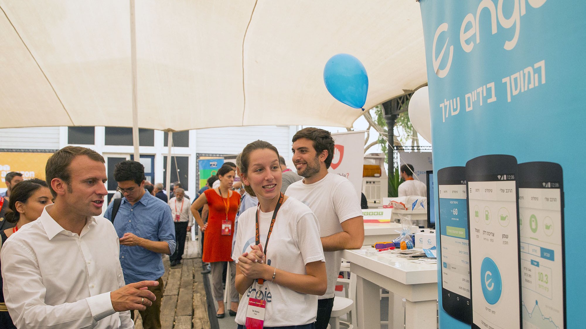 L'image montre une scène animée dans un événement, probablement un salon ou une foire. On peut voir des personnes interagissant autour d'un stand. Un homme, habillé en chemise blanche, semble discuter avec une jeune femme qui porte un uniforme avec un logo. À l'arrière-plan, il y a une bannière portant le logo d'Engie et des représentations d'applications sur des smartphones. L'ambiance est conviviale, avec des gens qui semblent engagés et intéressés par ce qui se passe.