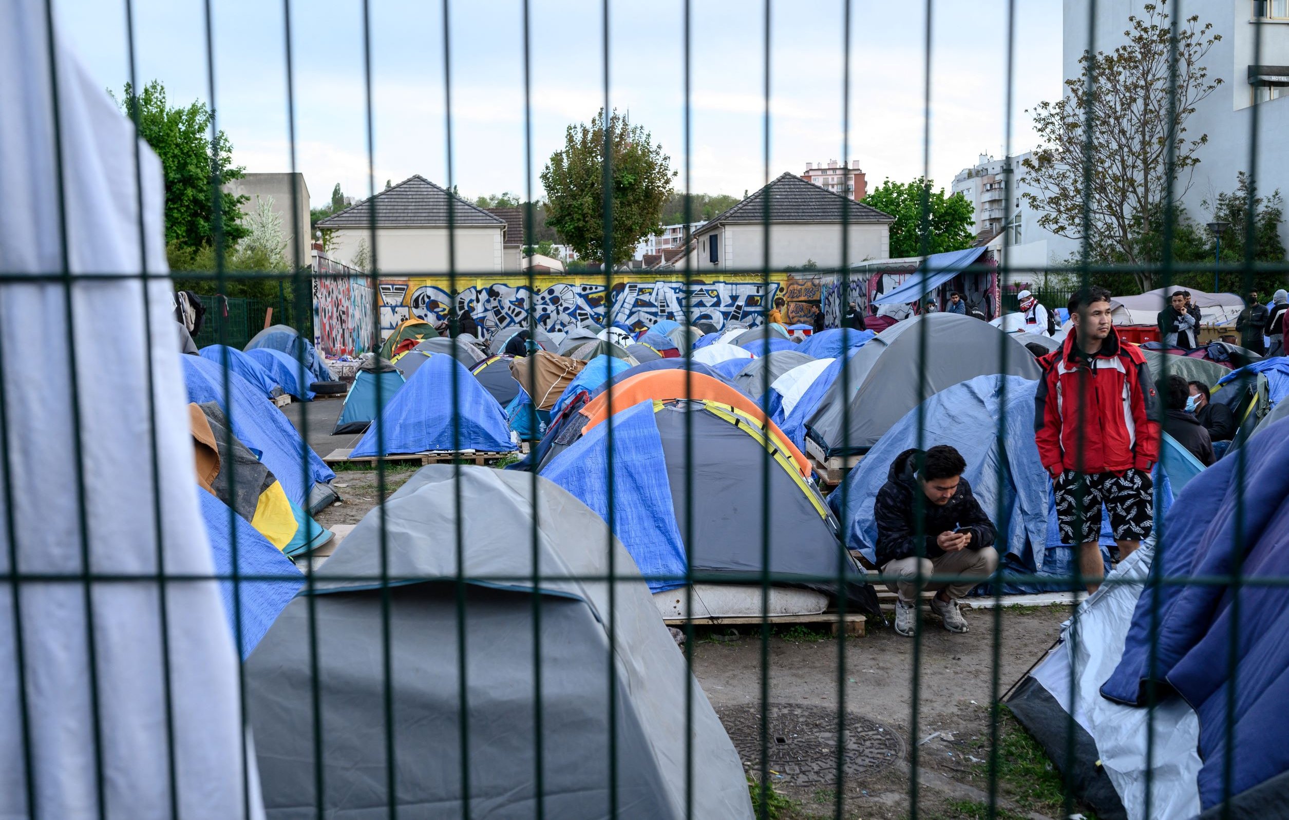 L'image montre un campement de tentes installé dans une zone urbaine. Les tentes sont de différentes tailles et couleurs, principalement en bleu et gris. On peut voir des personnes présentes, certaines debout et d'autres assises, semblant utiliser leur téléphone. En arrière-plan, il y a des bâtiments et un mur décoré de graffitis. L'ensemble du camp est entouré par une clôture, ce qui donne un aspect de confinement à la scène. Les arbres et la végétation ajoutent une touche de verdure au décor.