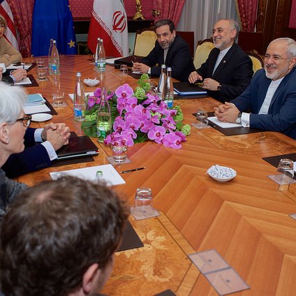 L'image montre une réunion diplomatique autour d'une grande table en bois. Des personnes sont assises de chaque côté, impliquées dans une discussion. On aperçoit des drapeaux d'États-Unis, de l'Union européenne et d'Iran sur la table. Au centre, il y a un bouquet de fleurs, probablement des orchidées. L'ambiance semble sérieuse mais productive, avec des participants au visage souriant, indiquant un échange positif.