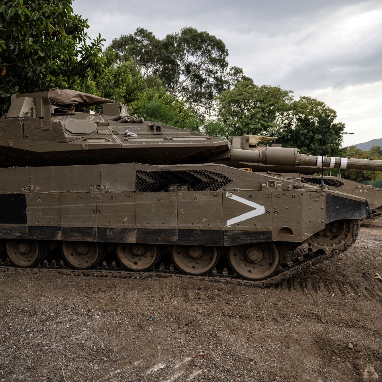 این تصویر یک تانک را نشان می‌دهد که در حالت ایستاده در کنار درختان قرار دارد. تانک دارای طراحی نظامی است و رنگ آن عمدتاً سبز زیتونی می‌باشد. در سمت چپ، شماره ۱۴ روی بدنه تانک دیده می‌شود. تانک به طور کلی ظاهری قوی و محکم دارد و می‌توان آن را در محیطی عمومی یا نظامی مشاهده کرد. زمین زیر تانک خاکی و خشک به نظر می‌رسد و آسمان کمی ابرناک است.