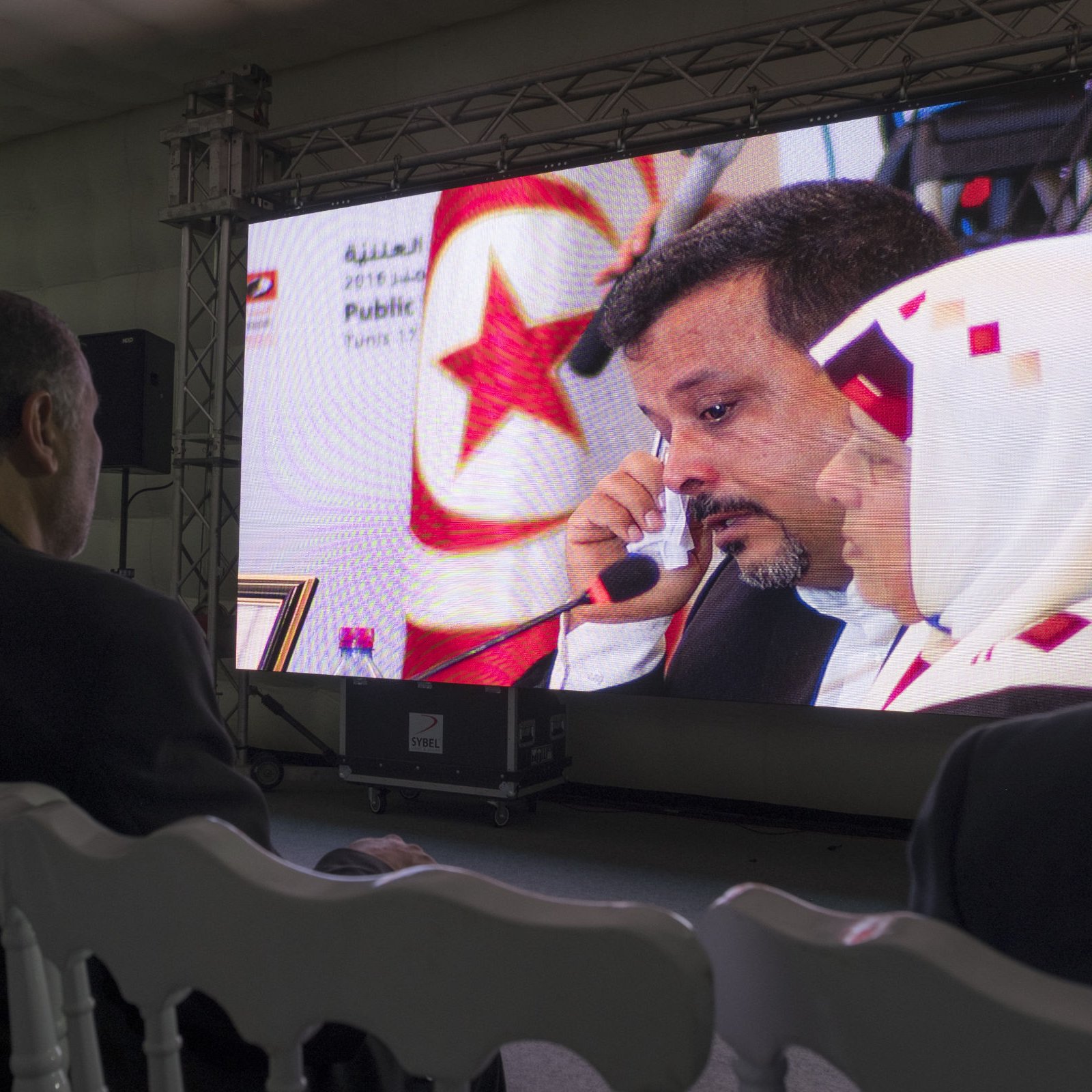 L'image montre un homme assis de dos, regardant un grand écran. Sur l'écran, on peut voir un homme parlant, avec une femme à ses côtés. L'ambiance semble formelle, peut-être liée à un événement officiel ou une conférence. L'éclairage et le décor sont modernes, avec des panneaux lumineux et des équipements audiovisuels visibles en arrière-plan. La scène dégage une atmosphère de concentration et d'attention.