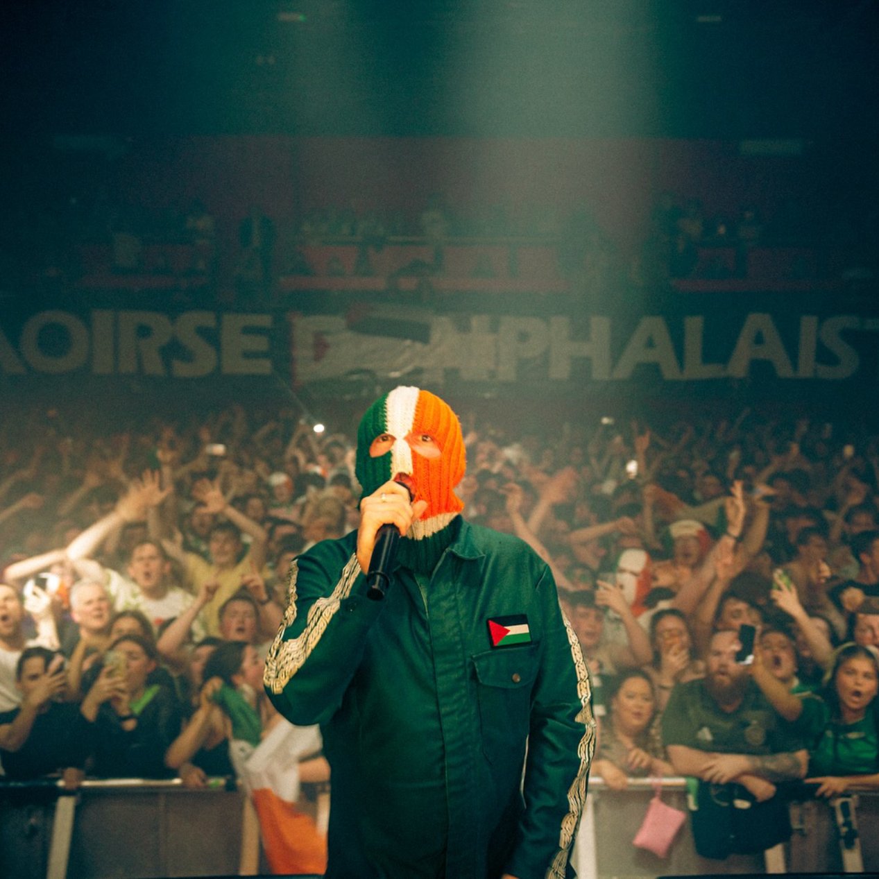 Un artiste en concert, masqué, devant une foule enthousiaste, avec des drapeaux irlandais et palestiniens.