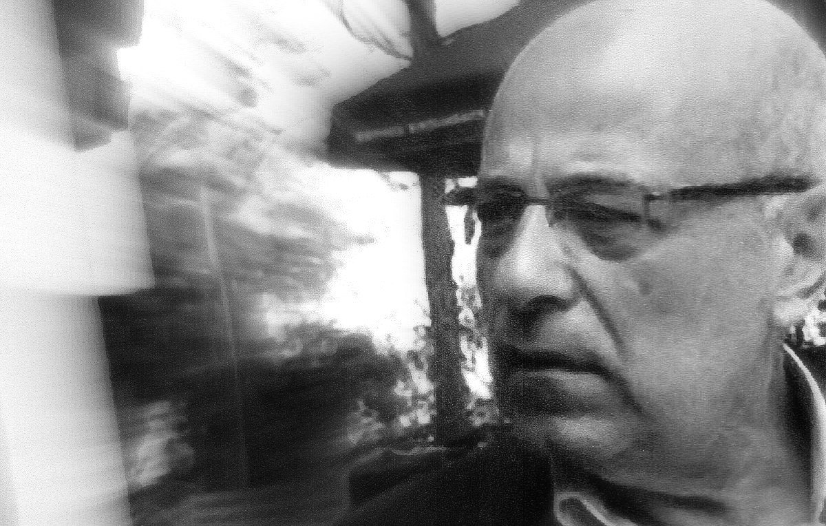 L'image présente un homme d'âge mûr avec des lunettes, légèrement flou en raison d'un effet de mouvement. Il semble pensif ou préoccupé, et son expression faciale transmet une certaine intensité. L'arrière-plan est indistinct, ce qui ajoute à l'effet dynamique de la photo. Les tonalités en noir et blanc renforcent le caractère dramatique de l'image.