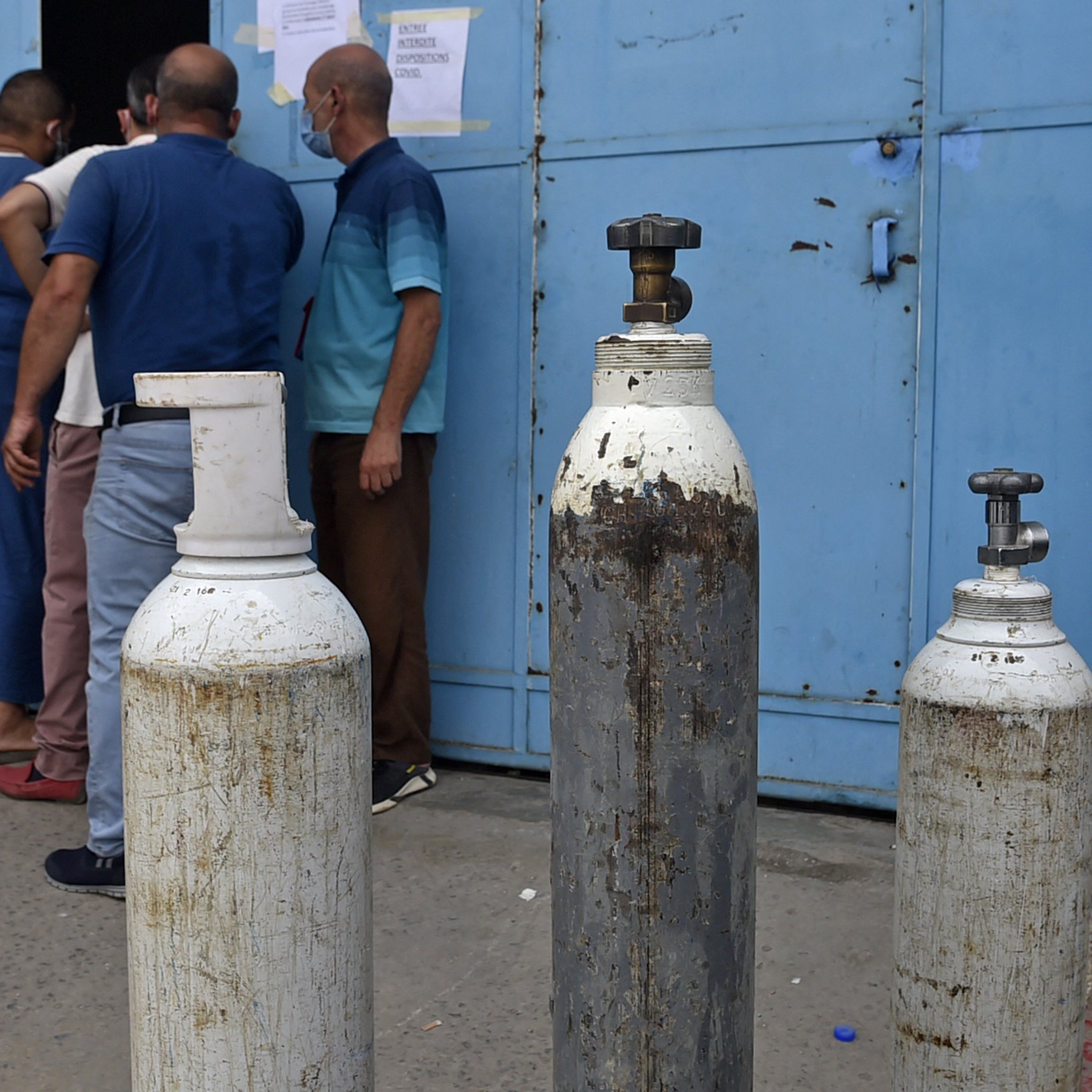 تظهر الصورة مجموعة من أسطوانات الغاز التي تُستخدم عادةً في المستشفيات أو للأغراض الصناعية. توجد ثلاث أسطوانات بأحجام متفاوتة، وتبدو واحدة منها قديمة ومُهملة. في الخلفية، يمكن رؤية بعض الأشخاص يقفون بالقرب من باب مبلوطة باللون الأزرق، مما يشير إلى موقع ربما يكون مركزًا لتوزيع الغاز أو خدمة طبية. تبدو الأجواء حيّة مع تواجد الأشخاص، مما يُبرز أهمية تلك الأسطوانات في الحياة اليومية.