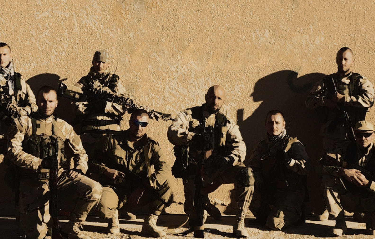 La imagen muestra a un grupo de soldados en un entorno desértico. Estos hombres están vestidos con trajes de combate y llevan equipamiento militar, incluyendo armas. Están organizados en una formación, posando para la foto. Algunos tienen gafas de sol y están en una posición que sugiere preparación y unidad. El fondo es simple, con una pared de color claro que resalta a los soldados.