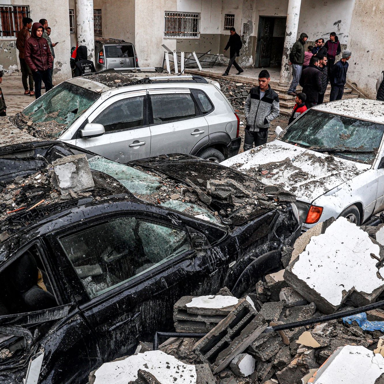 L'image montre des voitures endommagées, écrasées sous des débris dans une zone qui semble avoir subi un tremblement de terre ou un effondrement. Plusieurs personnes sont présentes, certaines observant la scène, d'autres apparemment en train de chercher ou d'évaluer les dégâts. L'environnement semble urbain, avec des bâtiments en arrière-plan, et l'atmosphère est chargée d'un sentiment de désastre et de désolation.