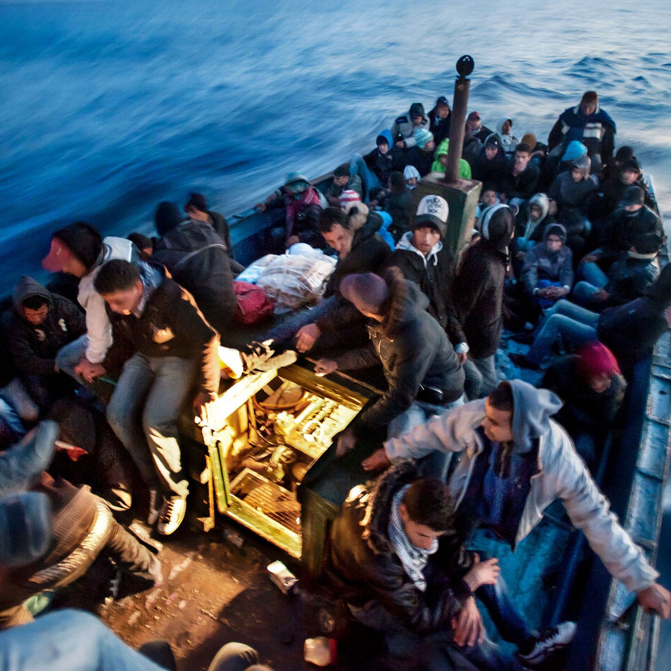 L'image montre une embarcation en mer, remplie de nombreuses personnes. Les passagers semblent être dans une situation difficile, tous étant assis ou debout, serrés les uns contre les autres. Ils portent des vêtements chauds, ce qui laisse supposer qu'ils font face à des conditions climatiques fraîches. L'eau est agitée et on peut voir des vagues, créant une atmosphère tendue et instable. La scène évoque des thèmes de migration et de recherche de refuge.