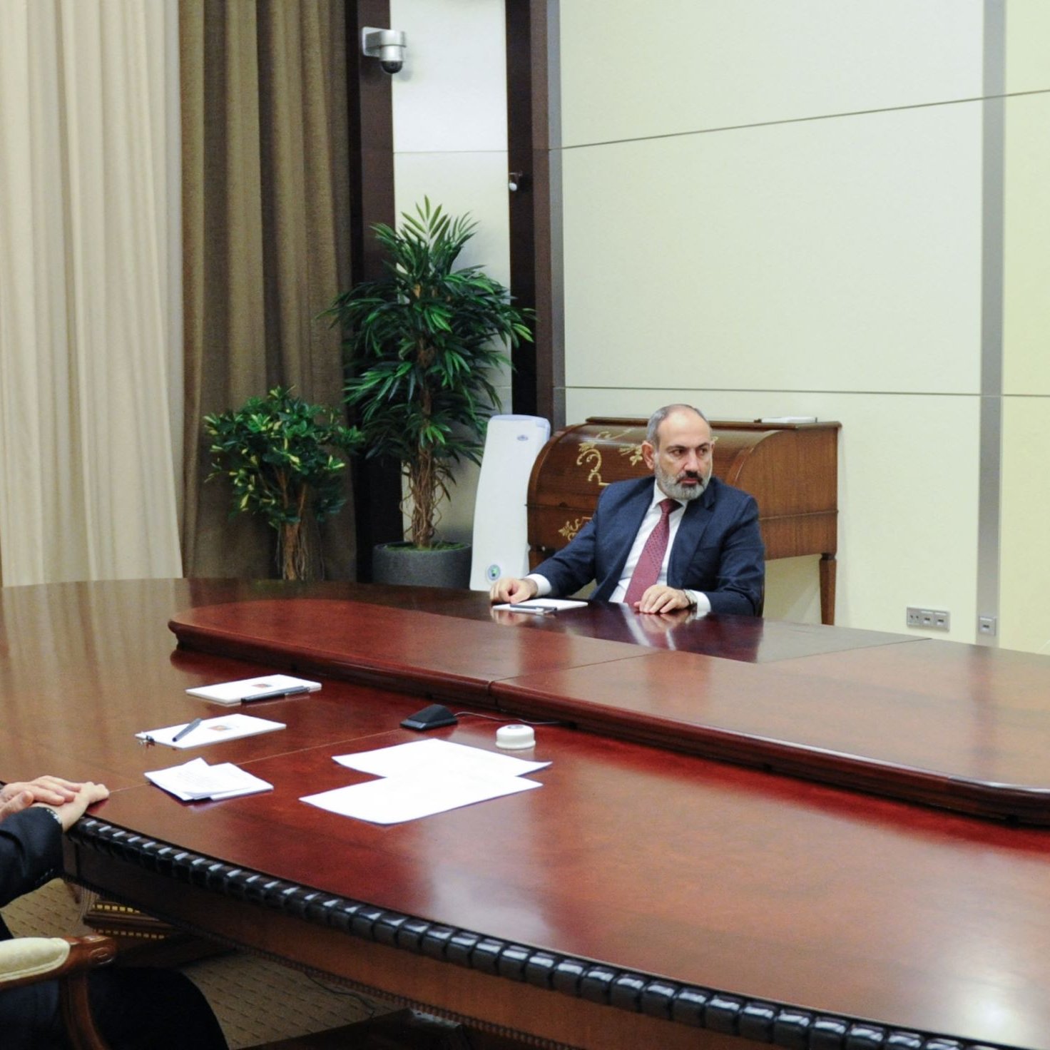 تصویر نشان‌دهنده یک جلسه رسمی است که در یک اتاق کنفرانس برگزار می‌شود. در مرکز میز بزرگ و قهوه‌ای رنگ سه نفر نشسته‌اند. روی میز تعدادی برگه و قلم دیده می‌شود. دیوارهای اتاق ساده و رنگ روشنی دارند و پرده‌های رنگ تیره در پشت افراد قرار دارد. در کنار اتاق، یک گیاه زینتی وجود دارد که فضایی طبیعی را به وجود می‌آورد. این تصویر به نظر می‌رسد مربوط به یک مذاکره یا نشست بین‌المللی باشد.