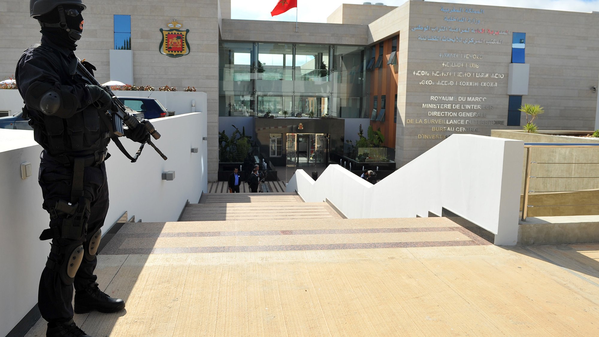 The image depicts a heavily armed police officer or security personnel standing at attention outside a building. The officer is dressed in black tactical gear and is holding a firearm. In the background, there is a modern structure with a large entrance, and the Moroccan flag is prominently displayed on top. The building appears to be a government facility, likely related to security or law enforcement, as indicated by the presence of the armed guard. The setting features stairs leading up to the entrance, with a clean, contemporary design.