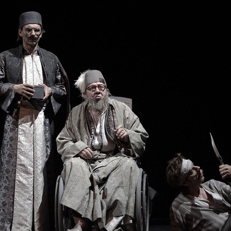 L'image représente une scène de théâtre mettant en avant trois personnages. Deux d'entre eux, habillés de façon traditionnelle et avec des vêtements sombres et ornés, semblent être en position d'autorité ou d'observation. Le troisième personnage est assis dans un fauteuil roulant, avec des vêtements plus clairs et un air pensif ou mélancolique. Il a une longue barbe grise et des lunettes. À côté de lui, un autre personnage, qui paraît être en détresse ou blessé, est au sol, tenant un objet pointu. L'éclairage est sombre, créant une atmosphère dramatique et intense.