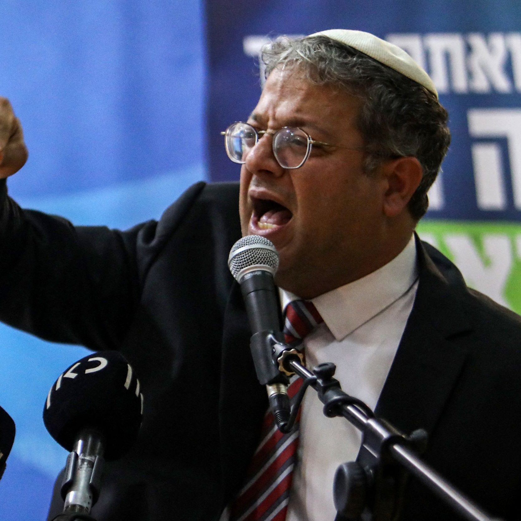 Nell'immagine si vede un uomo che parla appassionatamente al microfono, indossando un kippah. Sembra un politico o un oratore, con espressione intensa e gesti enfatici mentre esprime le sue idee. Sullo sfondo ci sono delle bandiere e scritte che potrebbero indicare un evento politico. L'atmosfera appare energica e coinvolgente.