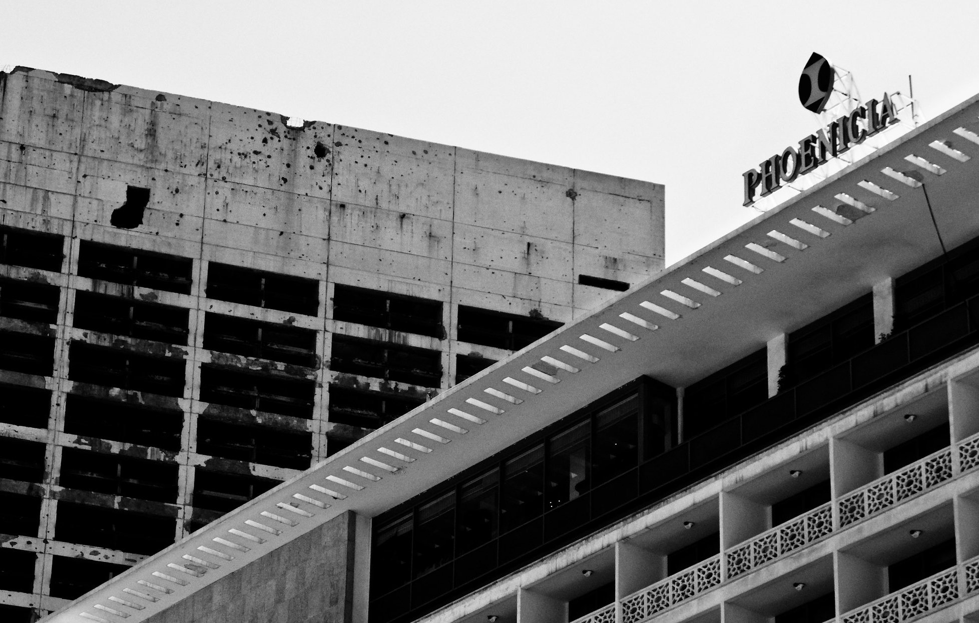 L'image montre deux bâtiments juxtaposés. À gauche, un bâtiment en béton brut et partiellement endommagé, avec des taches sombres visibles sur sa surface. À droite, un bâtiment plus moderne avec des lignes épurées et des balcons, arborant une façade blanche et des détails décoratifs. En haut à droite, le nom "Phoenix" est visible, probablement sur un panneau. L'ensemble de l'image est en noir et blanc, ce qui accentue le contraste entre les deux structures et leur état.