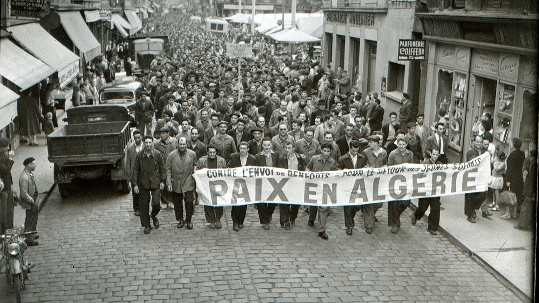 L'image montre une grande foule rassemblée dans une rue, probablement lors d'une manifestation. Les personnes marchent en portant une grande banderole qui indique "PAIX EN ALGÉRIE". On peut voir des hommes et quelques femmes, habillés de manière vestimentaire typique de l'époque. Des voitures et des commerces sont visibles en arrière-plan, ce qui donne une idée du contexte urbain. L'atmosphère semble sérieuse, suggérant que la manifestation est organisée autour d'un sujet important, en l'occurrence la paix en Algérie.
