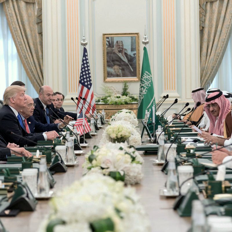 در این تصویر، یک جلسه رسمی در یک اتاق بزرگ برگزار می‌شود. در سمت چپ، تعدادی از مقام‌های آمریکایی نشسته‌اند و پرچم ایالات متحده در کنار آنها قرار دارد. در سمت راست، مقام‌های سعودی نشسته‌اند و پرچم عربستان سعودی به چشم می‌خورد. بر روی میز، گل‌های سفید و دیگر وسایل اداری وجود دارد. این جلسه نشان‌دهنده تعاملات دیپلماتیک و همکاری‌های بین‌المللی بین دو کشور است. دیوارهای اتاق با پرده‌های زیبا تزئین شده و تصویری بزرگ از یک شخصیت تاریخی در پس‌زمینه قرار دارد.