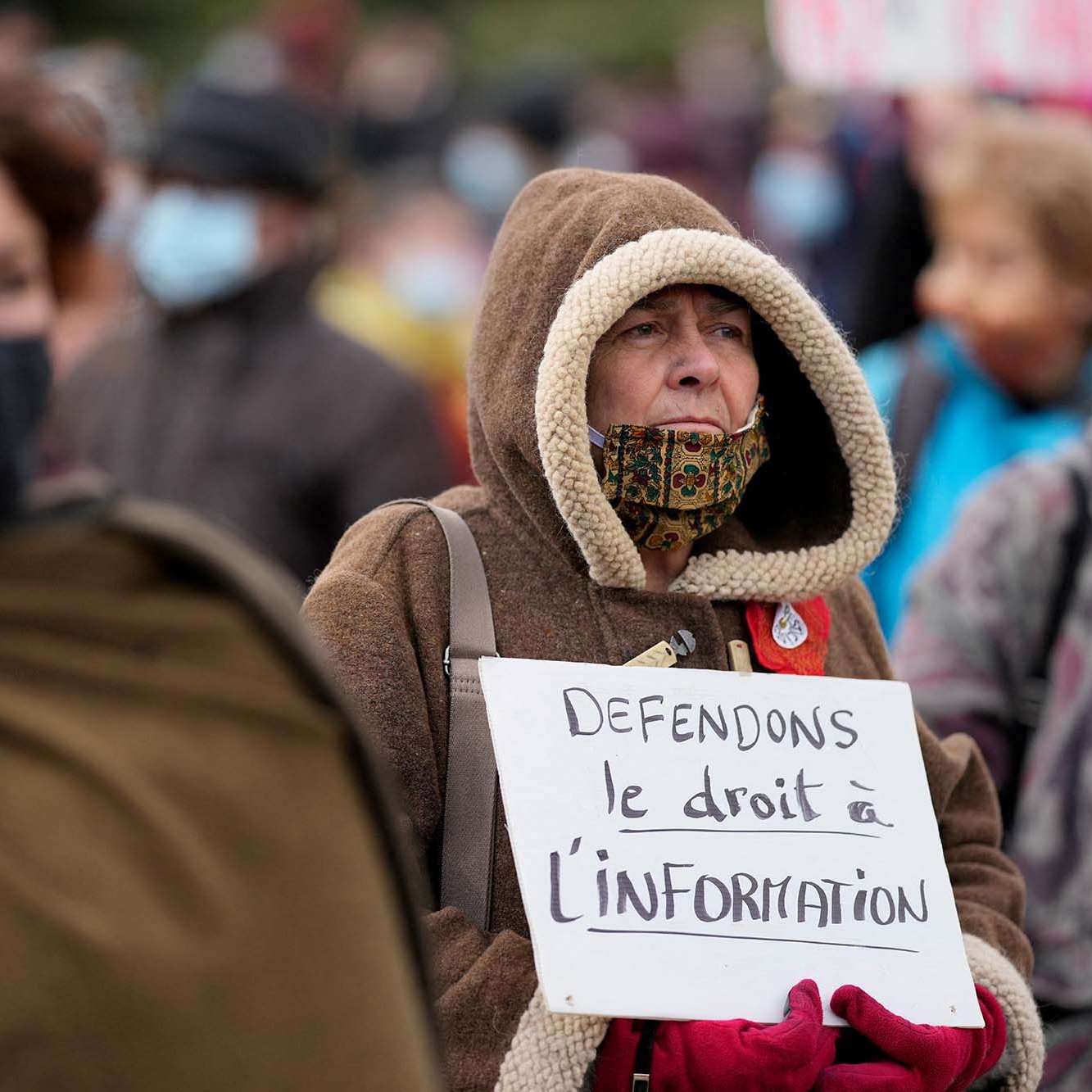 L'image montre un groupe de personnes rassemblées lors d'une manifestation. Au premier plan, une femme âgée porte une grande capuche et un masque, tenant une pancarte sur laquelle il est écrit : "Défendons le droit à l'information". Les autres manifestants sont flous en arrière-plan, mais on peut voir qu'ils portent également des masques. L'atmosphère semble sérieuse et engagée, avec des personnes qui militent pour leurs droits.